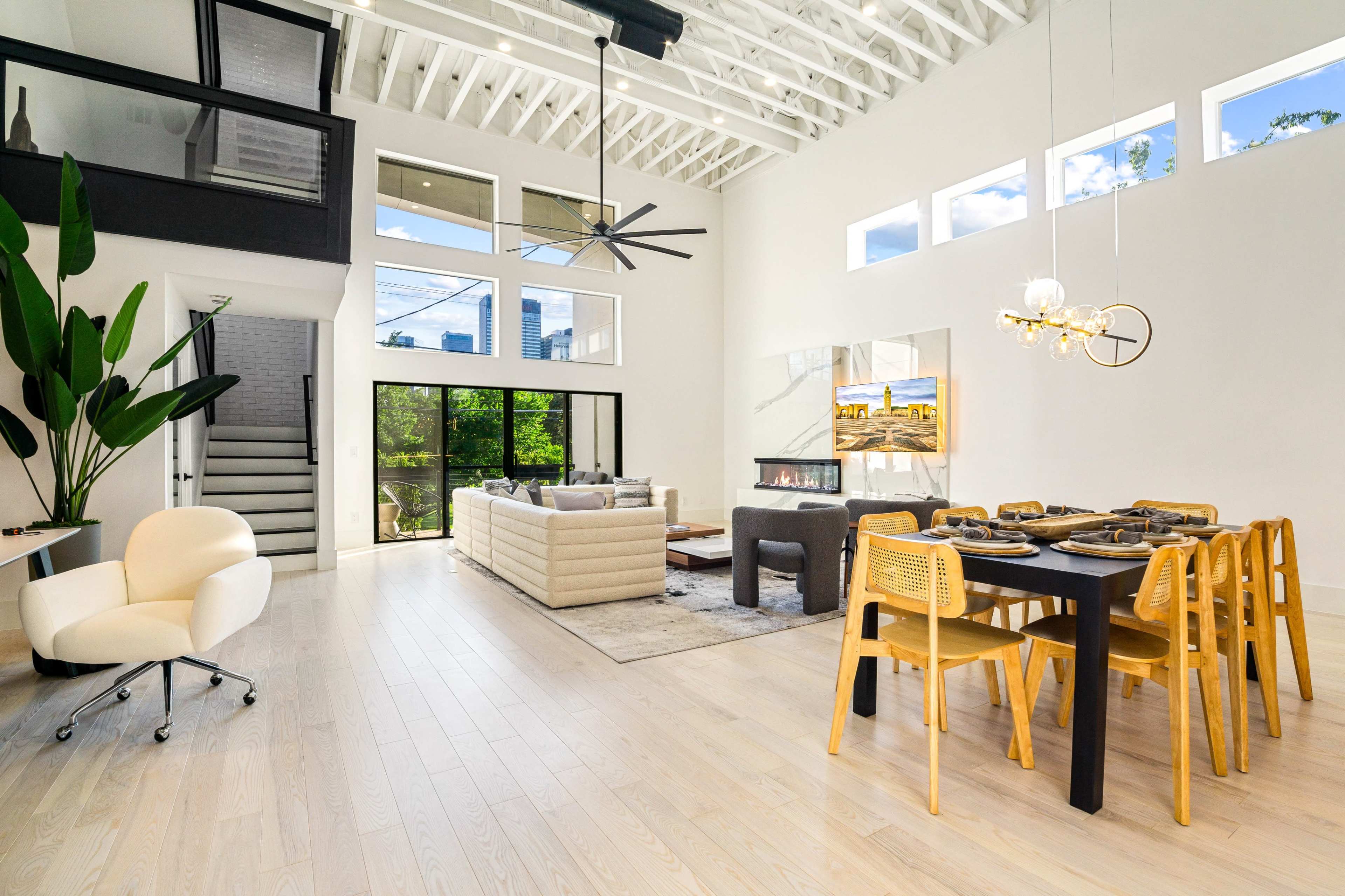 A modern living space with a dining area, plush seating, large windows, and a sleek staircase.