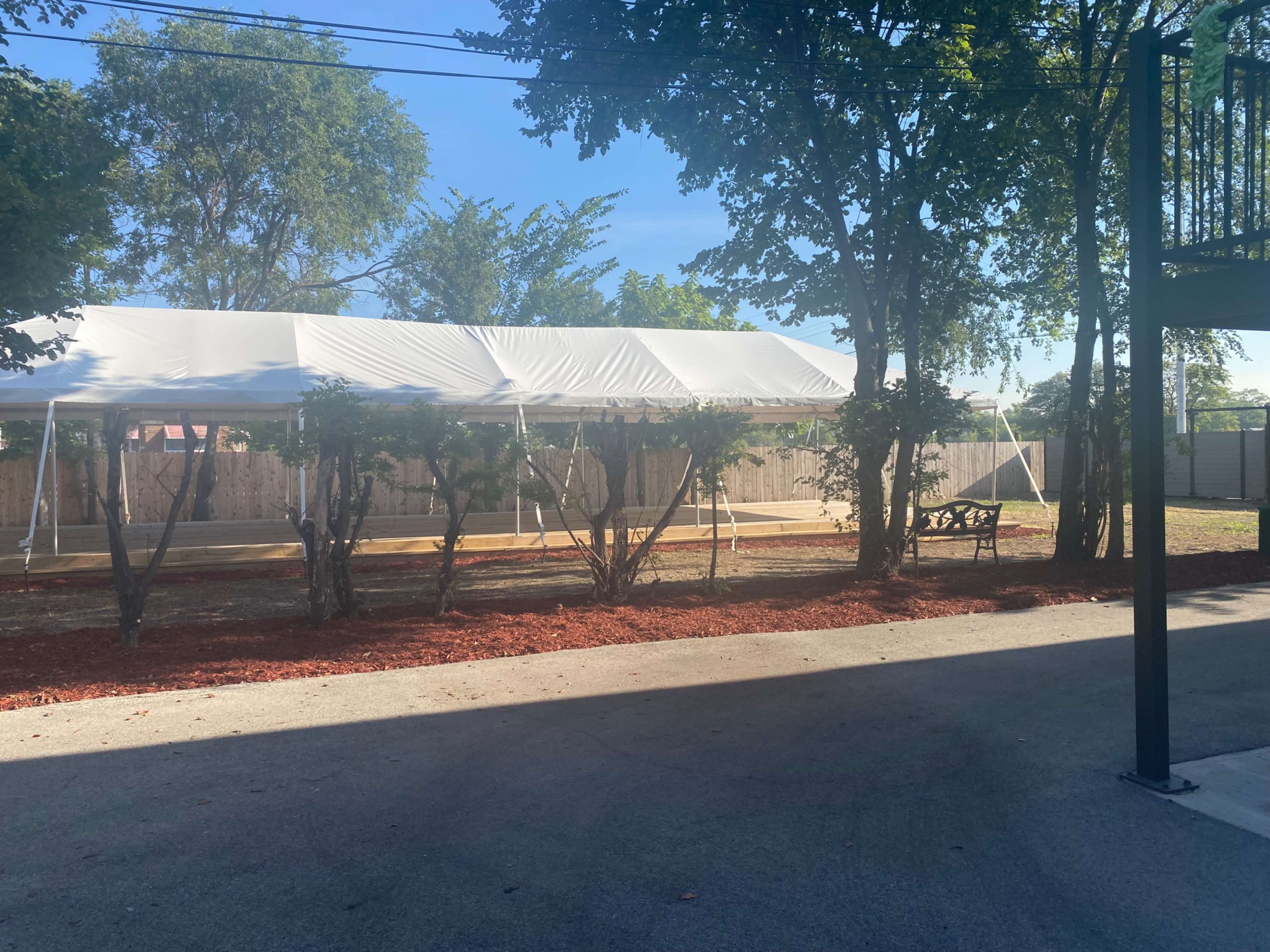 A large white tent is set up in a sunlit outdoor area surrounded by trees and freshly laid mulch.