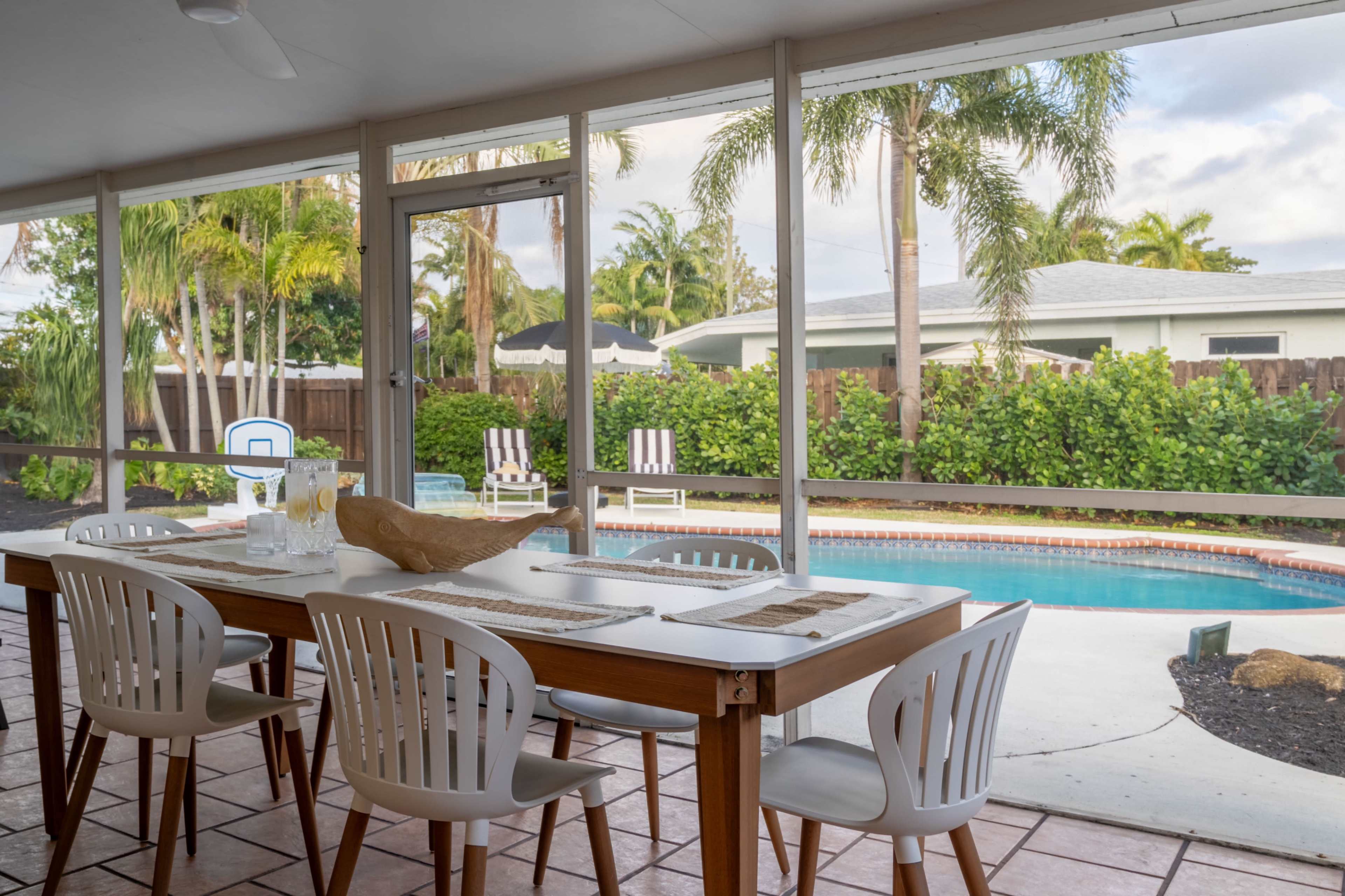 The image shows a dining table set on a patio with a view of a swimming pool and palm trees in the background.