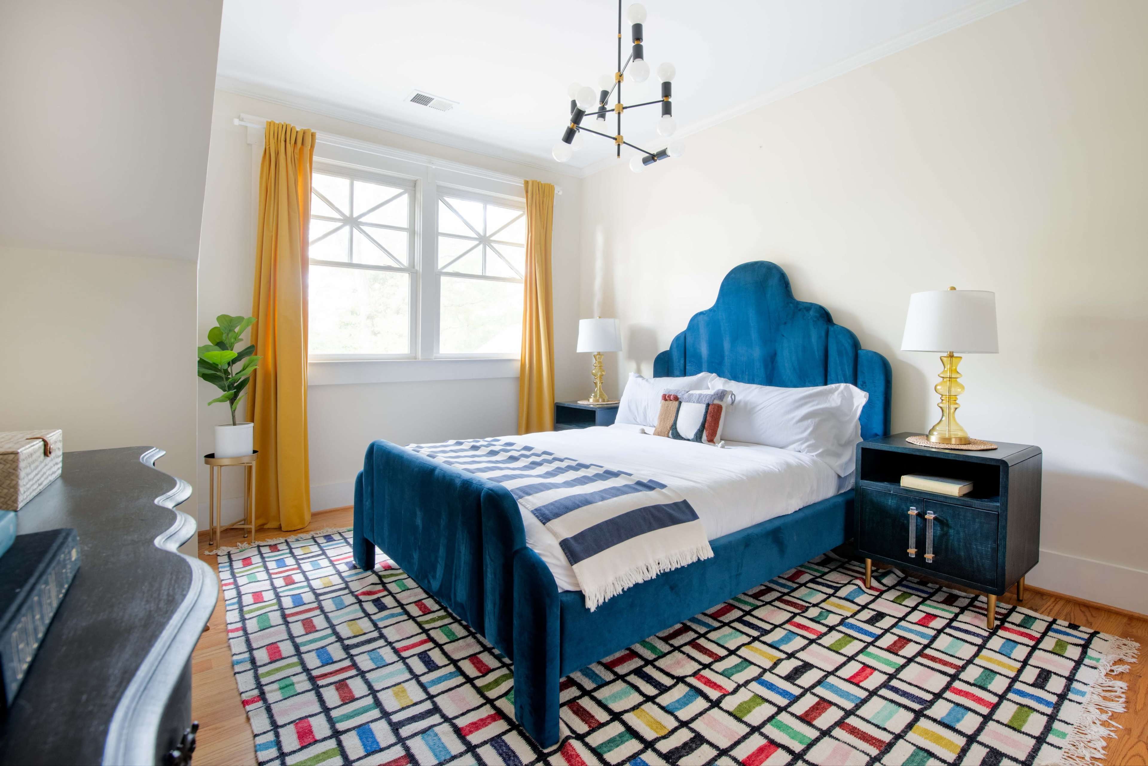 The image shows a stylish bedroom featuring a blue upholstered bed, a colorful patterned rug, white linens, yellow curtains, and two side tables with lamps.