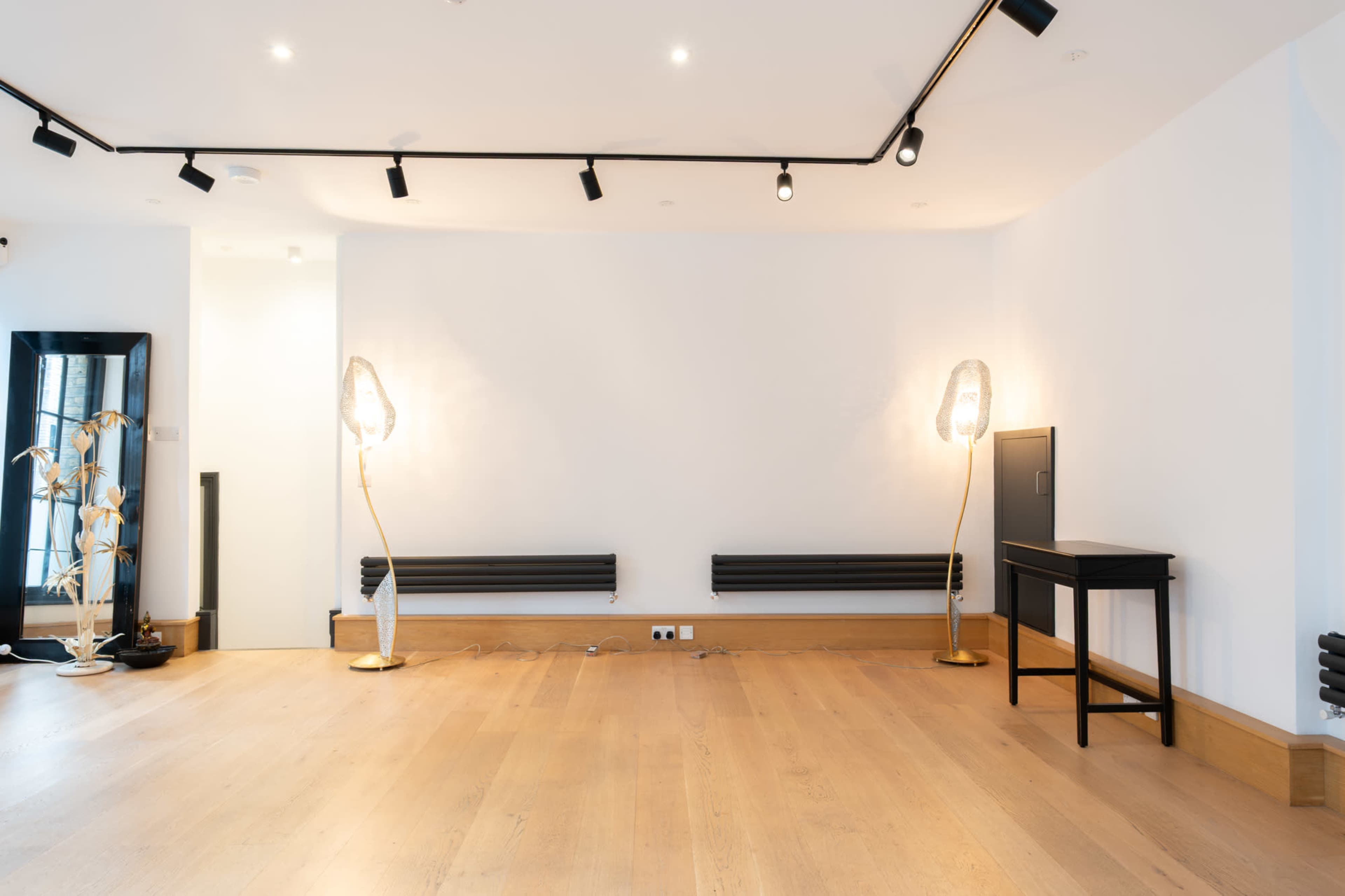 The image features a minimalist interior with wooden flooring, a small black table, two free-standing lamps, and a blank wall.