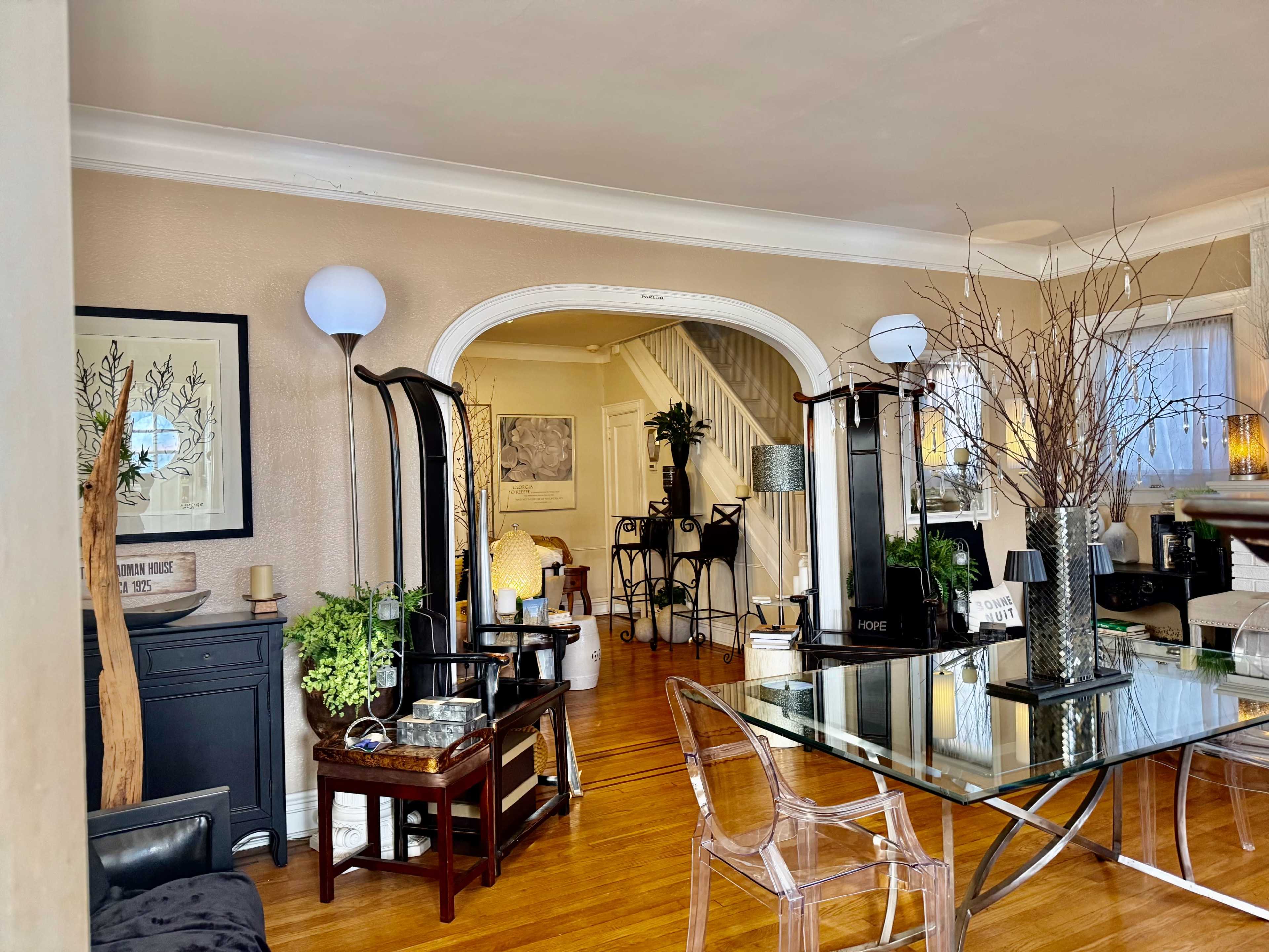 The image shows a spacious living room area with a glass table, modern furnishings, and decorative plants.