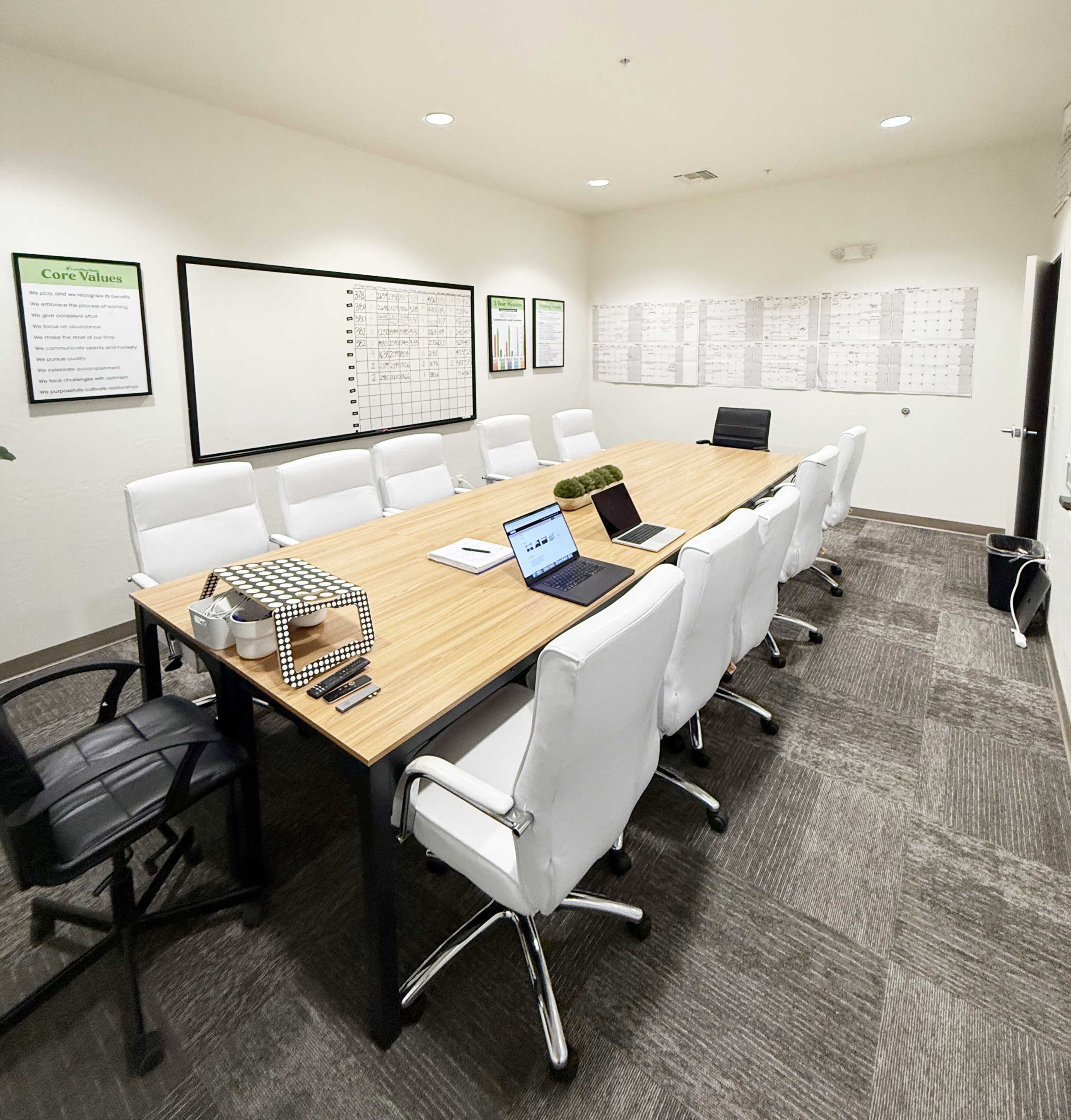 A large conference room features a long wooden table surrounded by white chairs, with a laptop and decorative items on the table, and charts displayed on the walls.