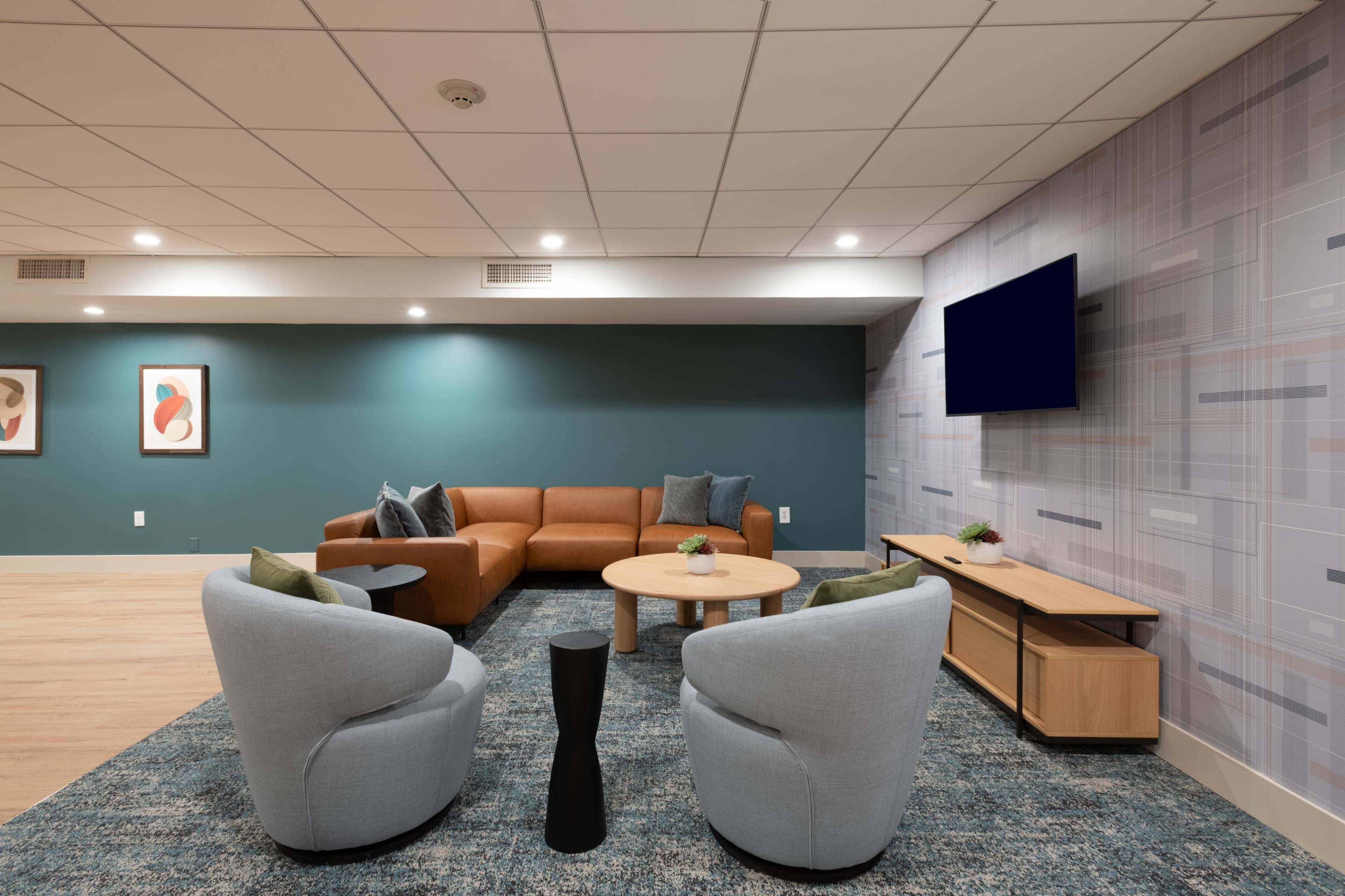 The image shows a modern lounge area featuring a brown sectional sofa, two light blue armchairs, a round wooden coffee table, and a television mounted on the wall.