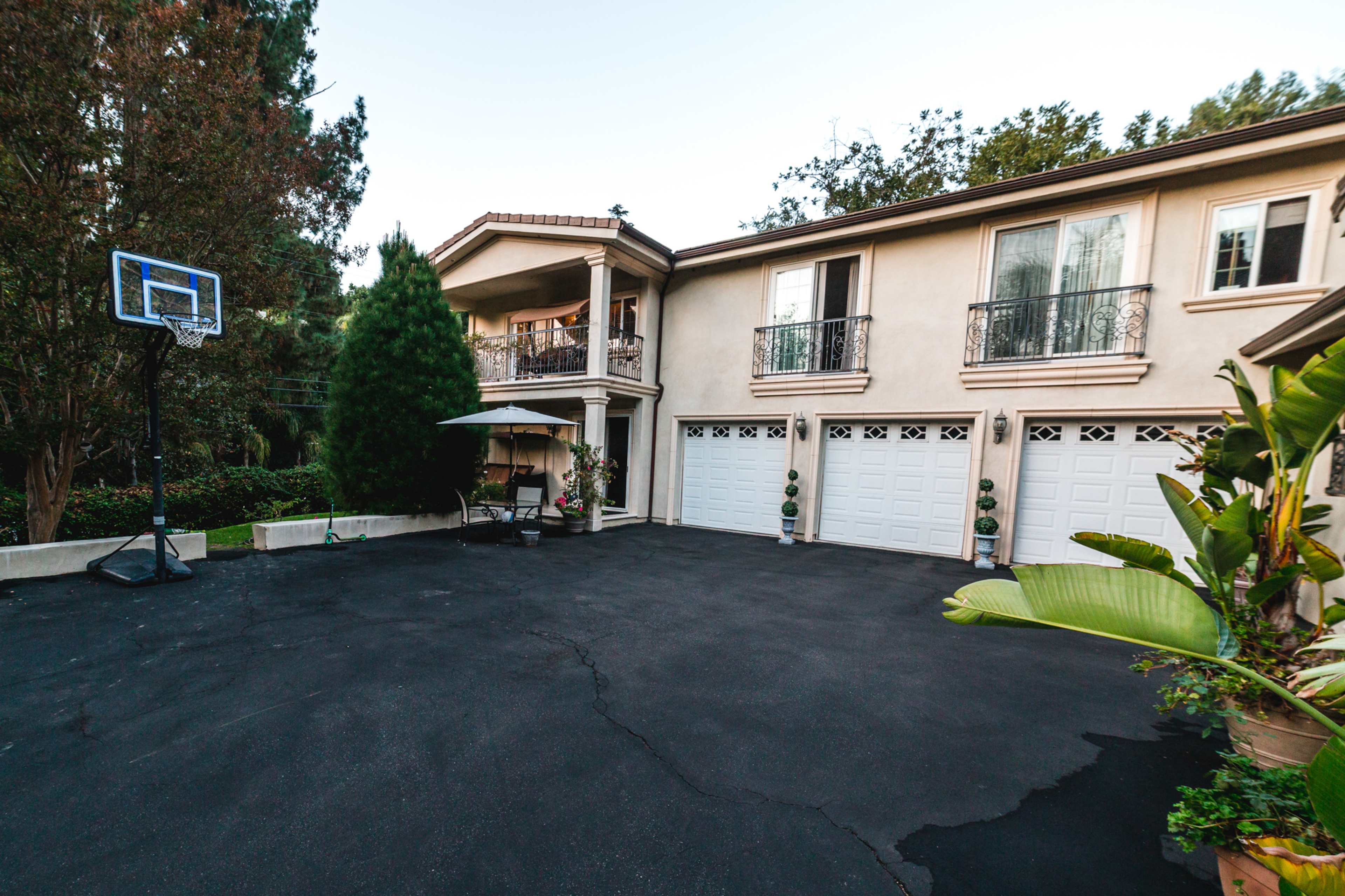 The image shows a large home with a basketball hoop in the driveway and a covered seating area amidst landscaped greenery.