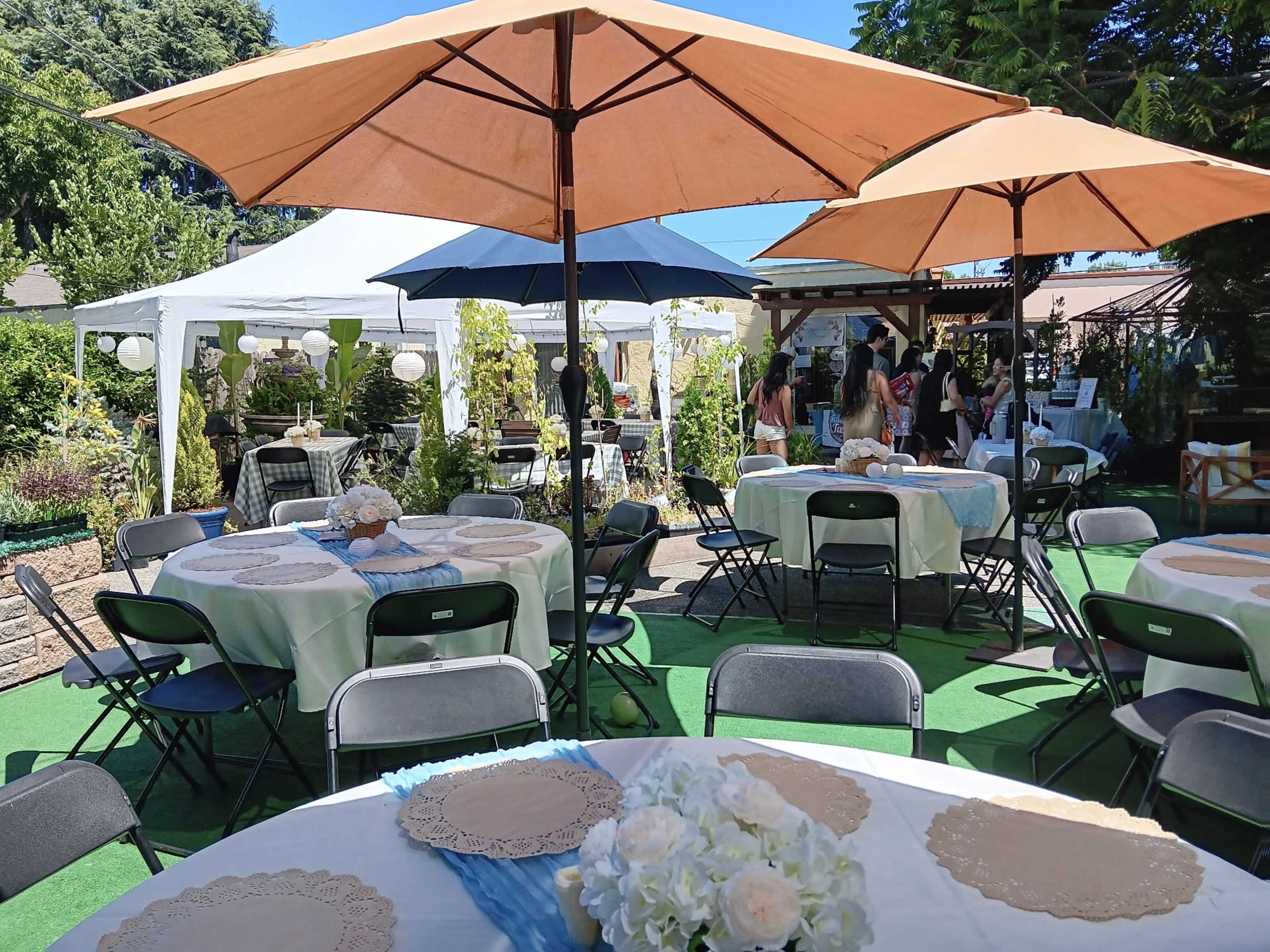 The scene shows an outdoor event setup with round tables under large umbrellas, surrounded by greenery and tents in the background.