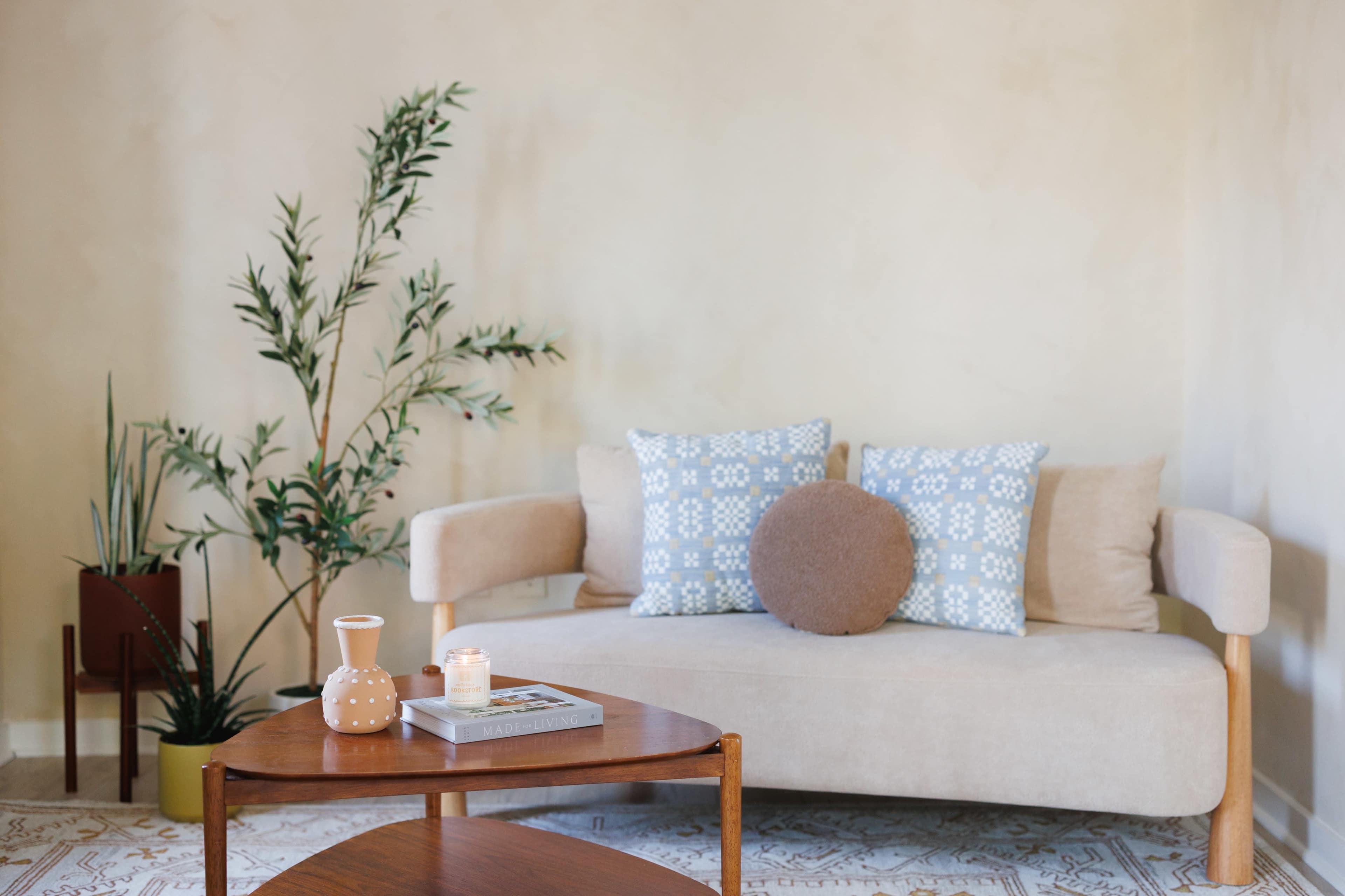 A light-colored couch with patterned cushions sits beside a small wooden table and a candle, while plants are positioned in the corner of the room.