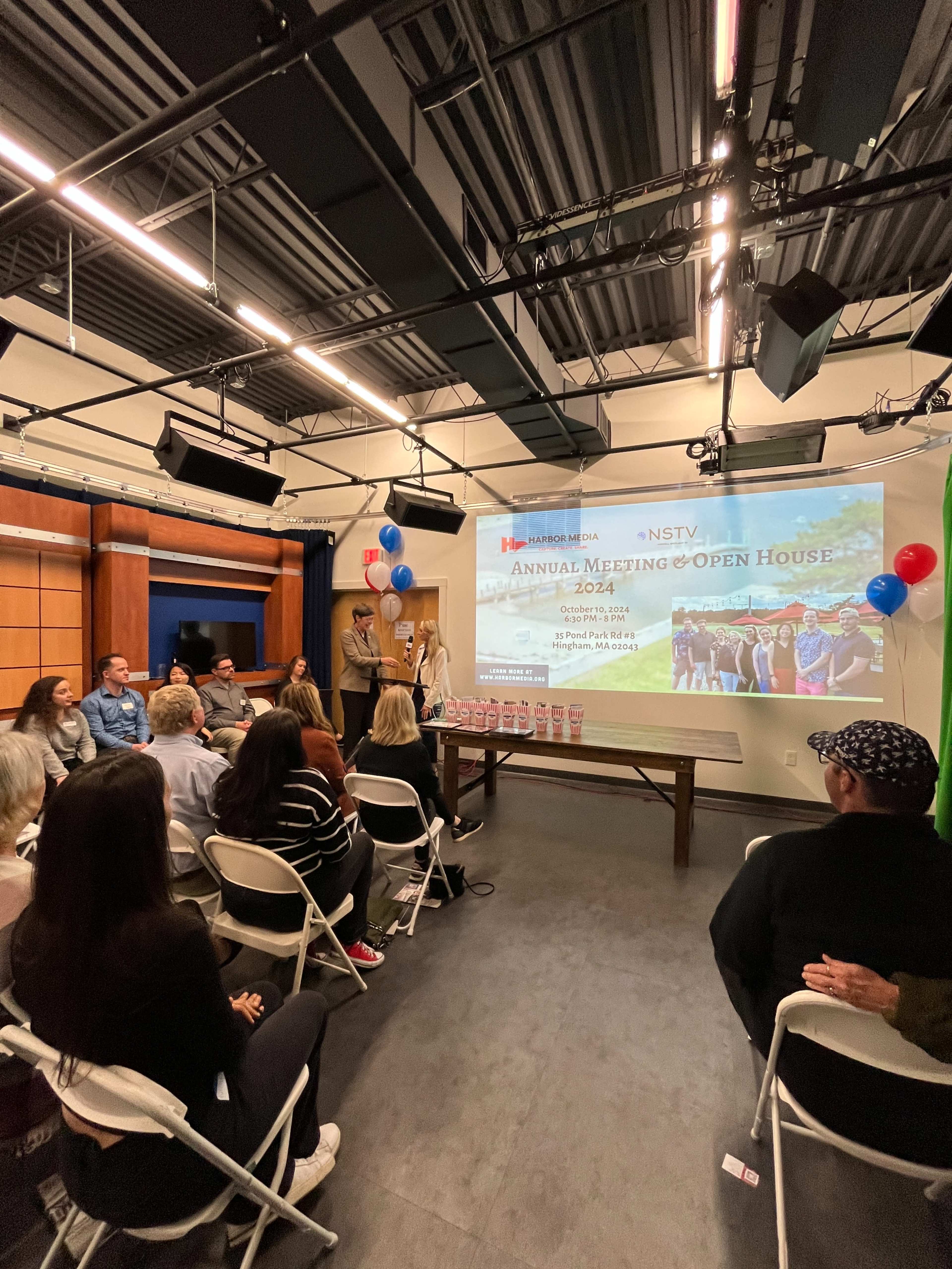 A group of people is seated in a studio, watching a presentation that is being delivered on a screen at the front, decorated with balloons for an annual meeting and open house event.