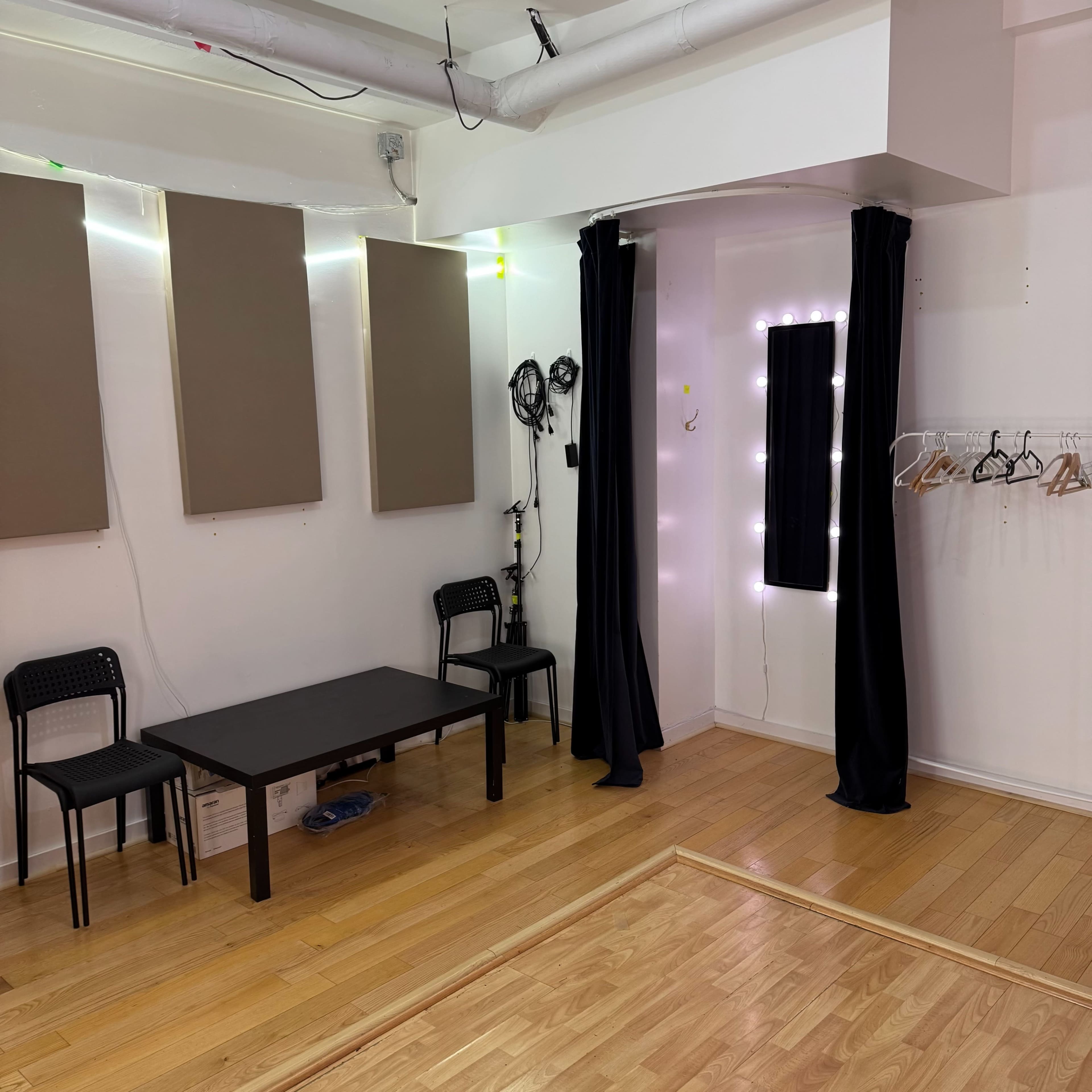 The image shows a small room with wooden flooring, containing a table, two chairs, and a dressing area with lights and curtains.