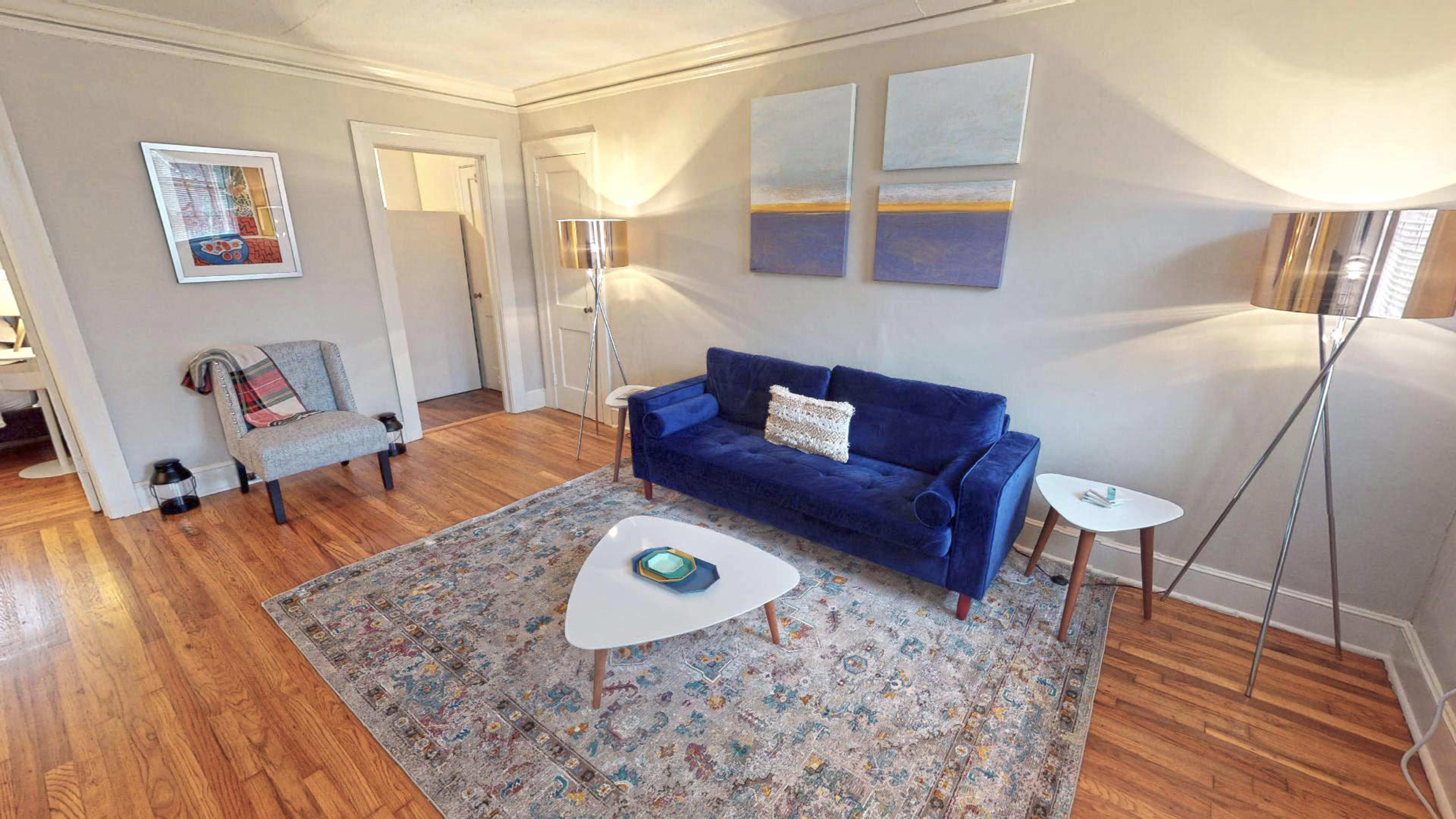 The image shows a living room featuring a blue sofa, two floor lamps, a small side table, and a patterned area rug on wooden flooring.