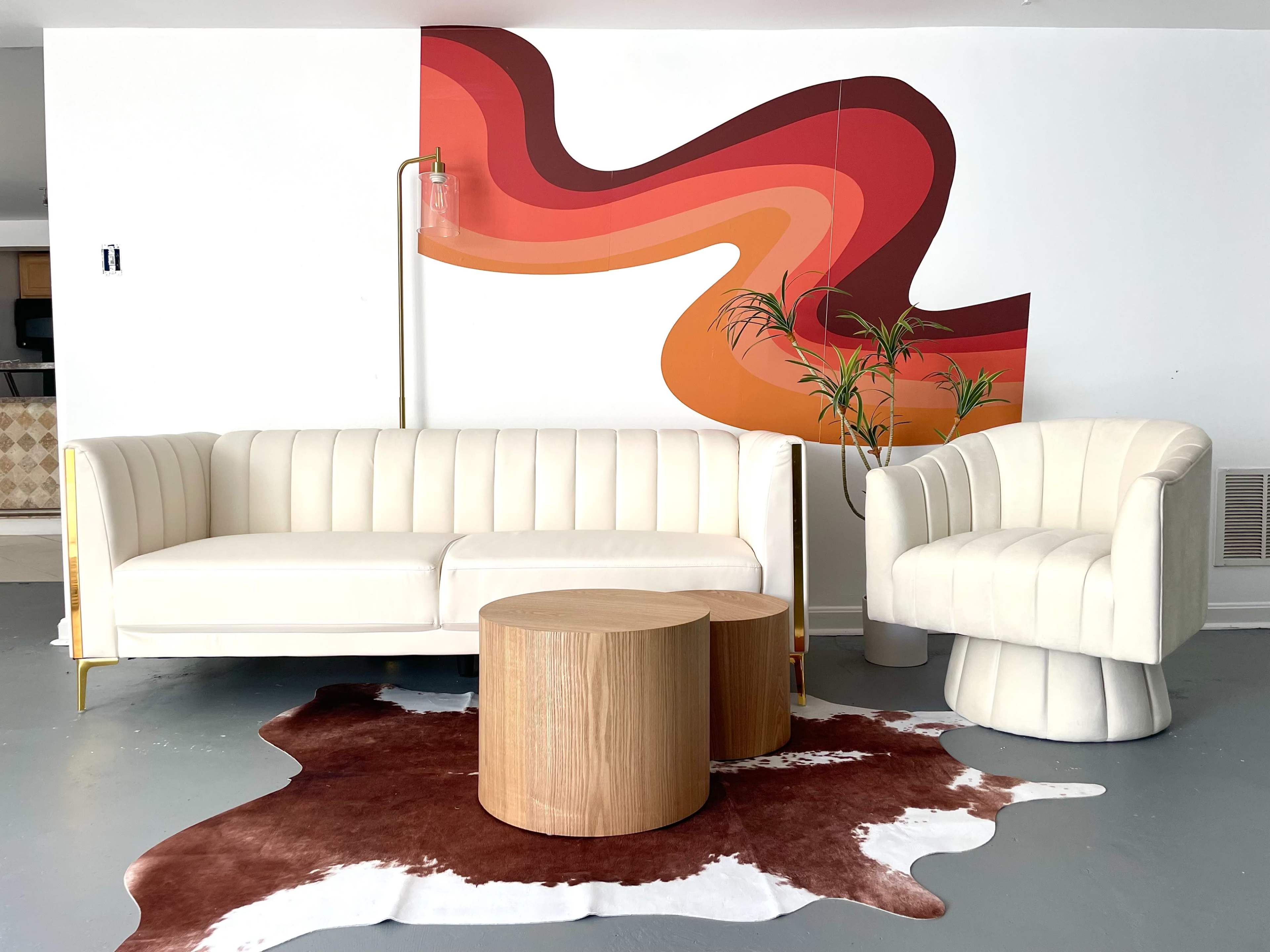 A modern living room setup with a white, ribbed sofa and armchair, a pair of wooden side tables, and a colorful abstract mural on the wall behind them.
