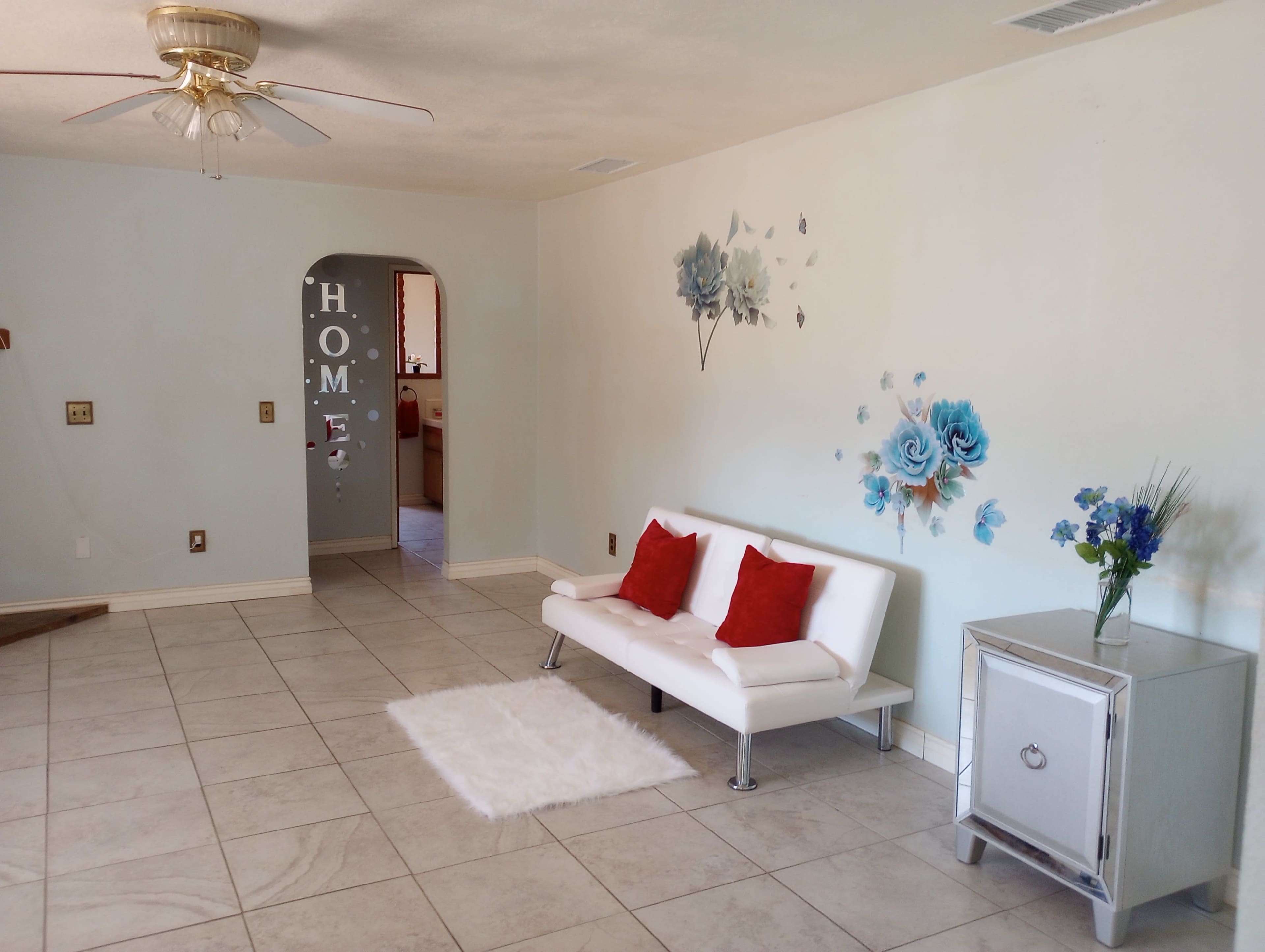 A living room with a white sofa adorned with red cushions, a small silver cabinet, and a floral wall decoration.