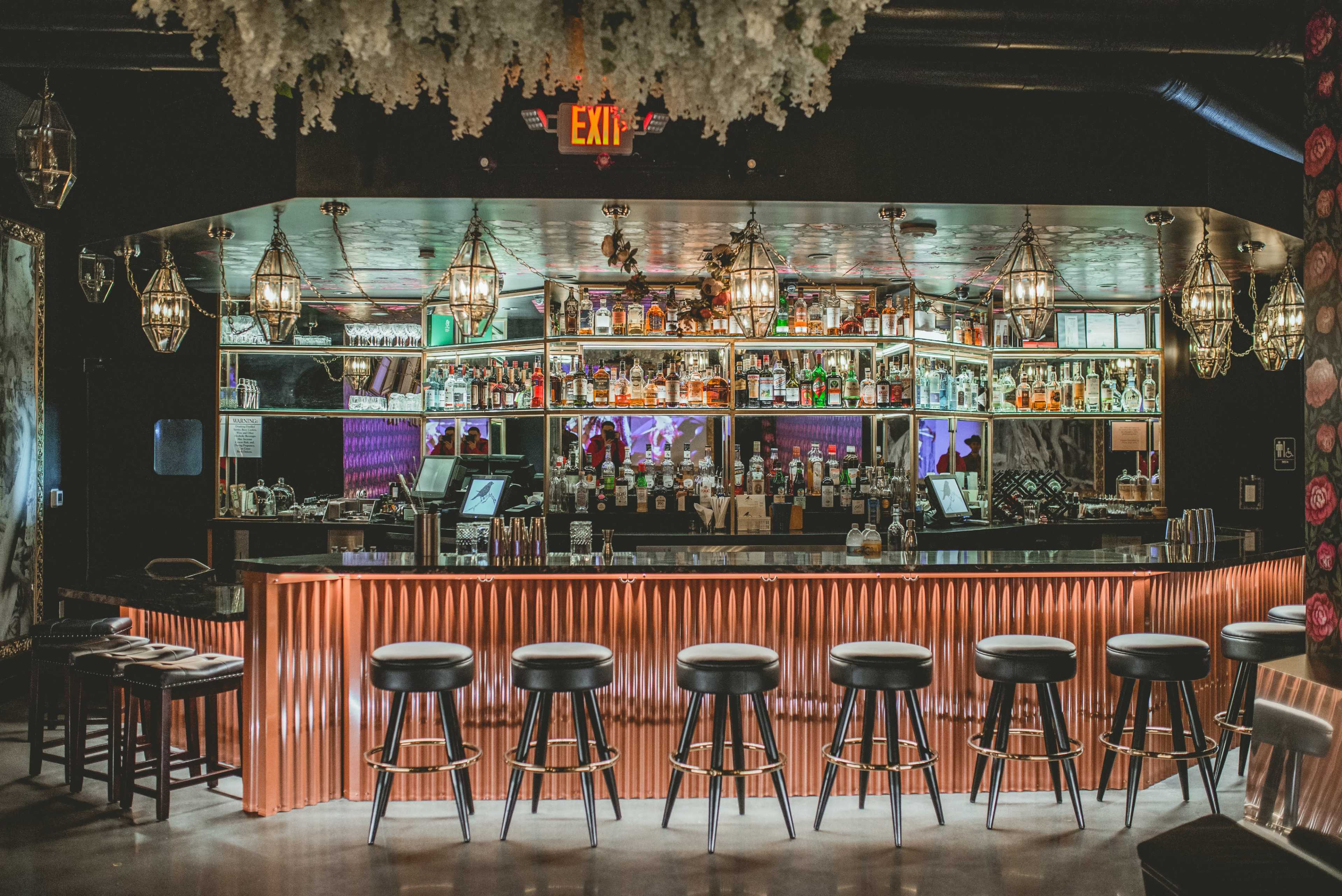 A well-lit bar with a polished countertop and several high-backed stools, showcasing an array of alcoholic beverages on shelves behind it.