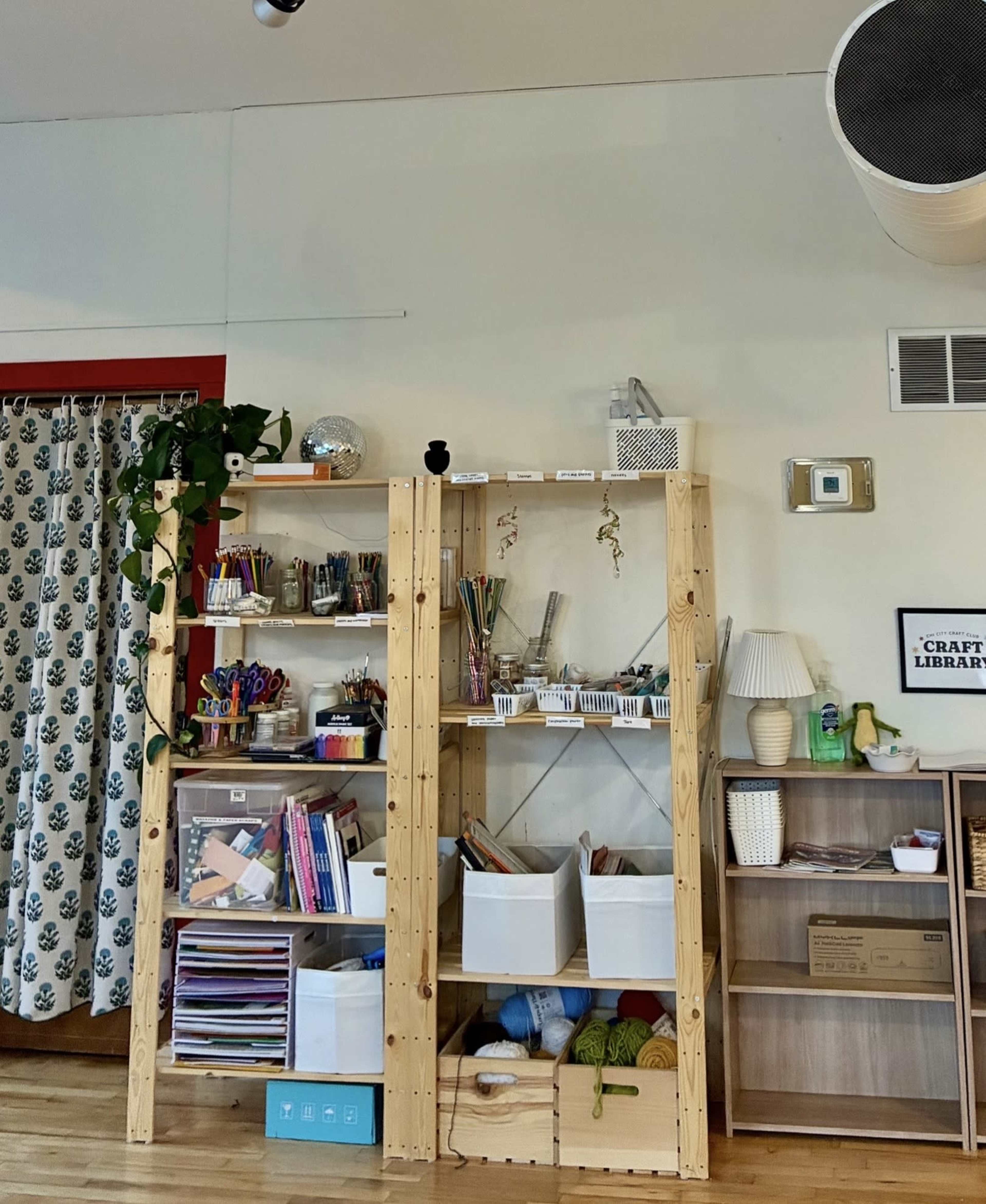 The image shows a well-organized craft space with a wooden shelving unit displaying various art supplies, books, and storage bins against a wall decorated with a patterned curtain.