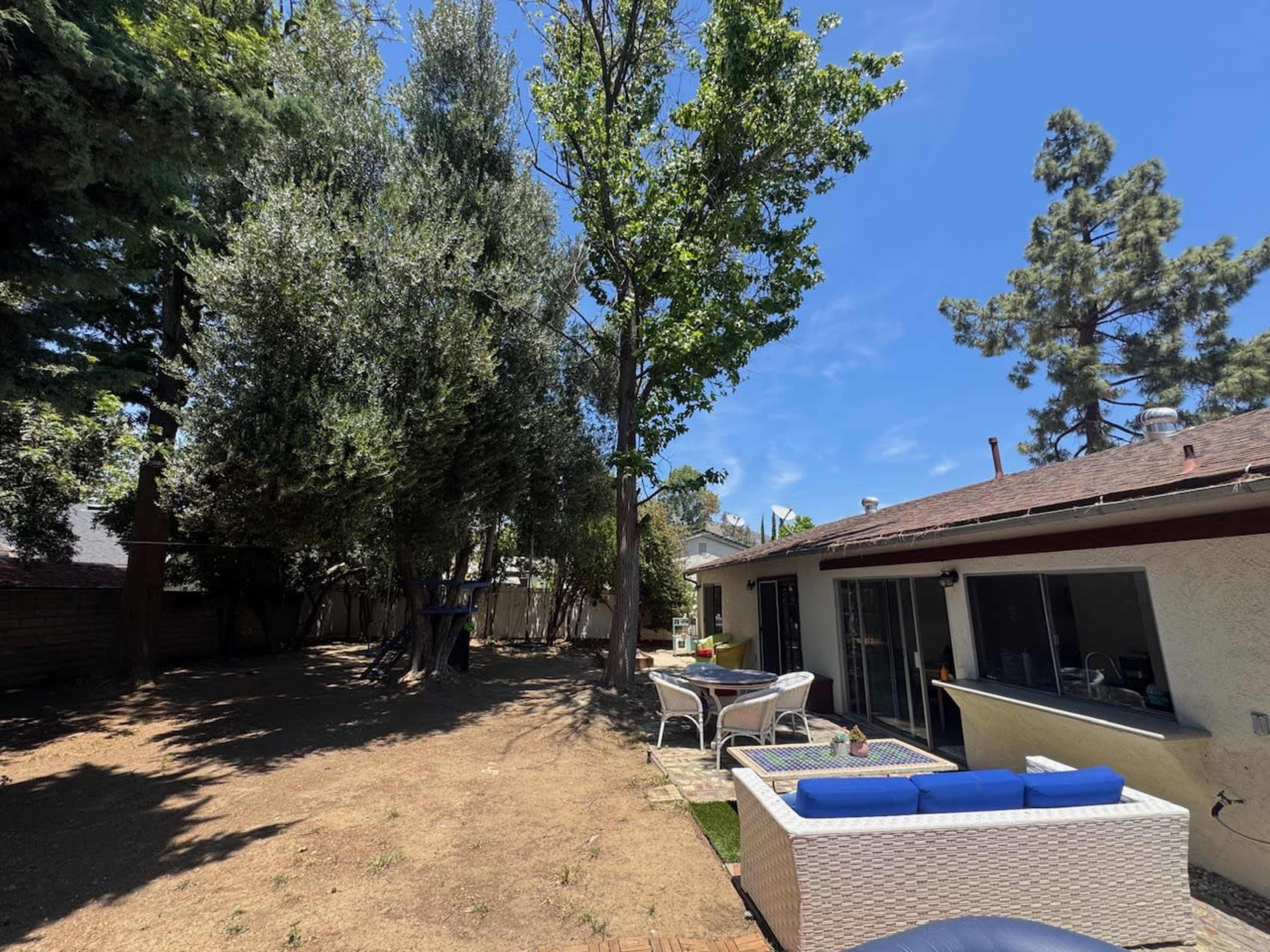 A backyard features sparse vegetation with a seating area in the foreground and a house on the right under a clear blue sky.