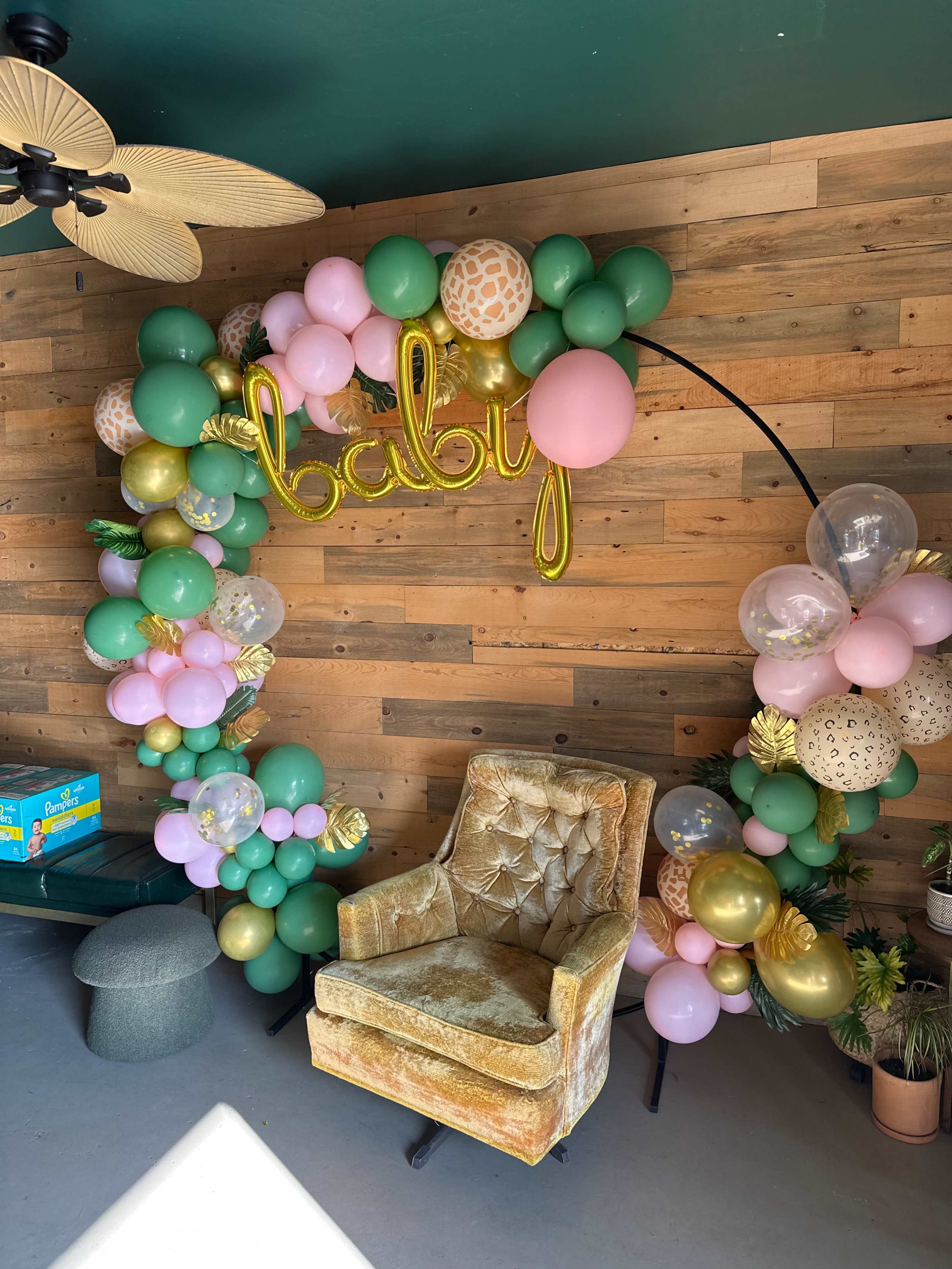 The image features a decorated corner showcasing a balloon arch in green, pink, and gold with the word "baby" in gold letters, alongside a vintage-style golden armchair.