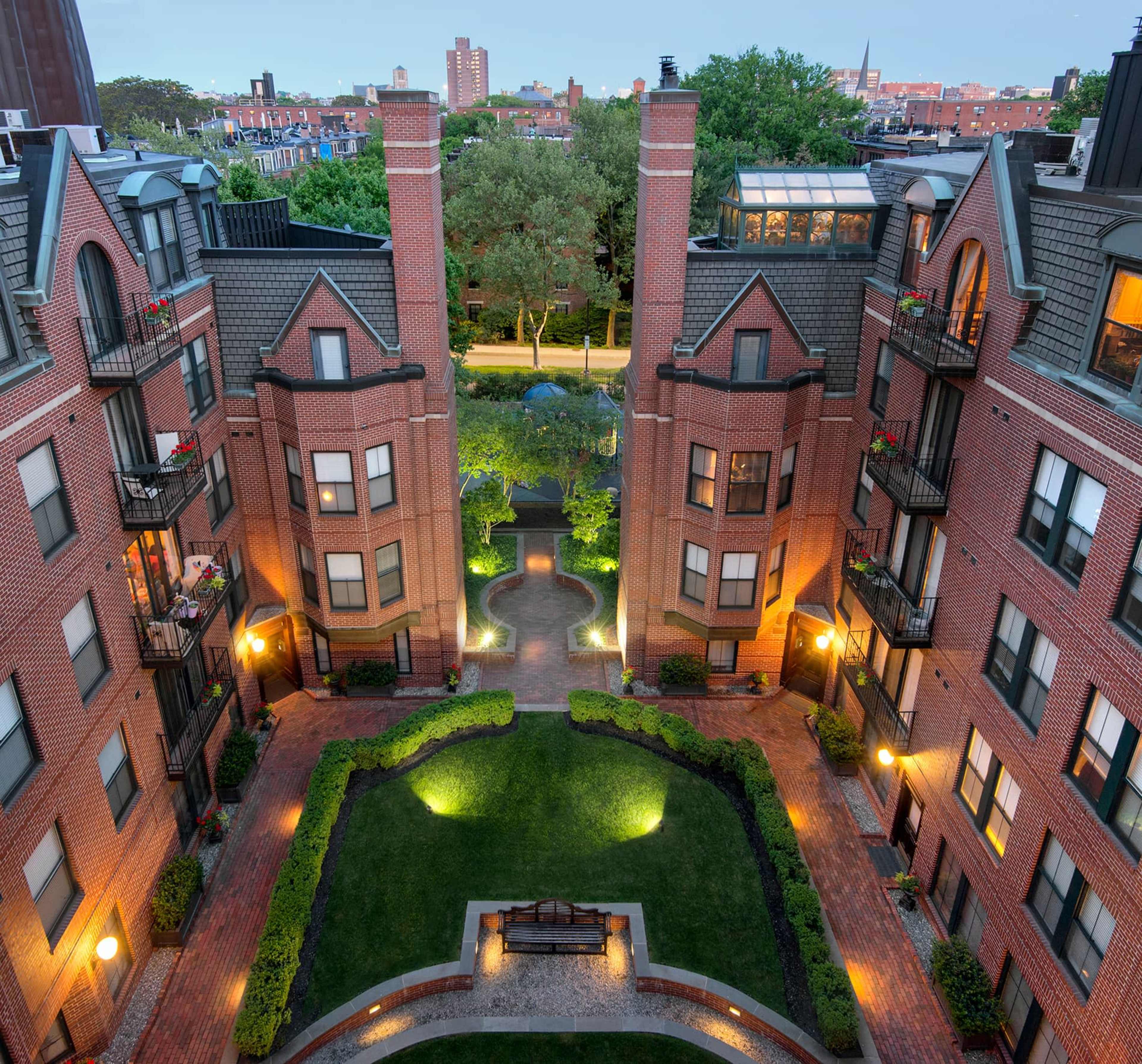 The image shows a landscaped courtyard surrounded by brick apartment buildings, with pathways lined by lights and trees, set against a backdrop of city buildings.