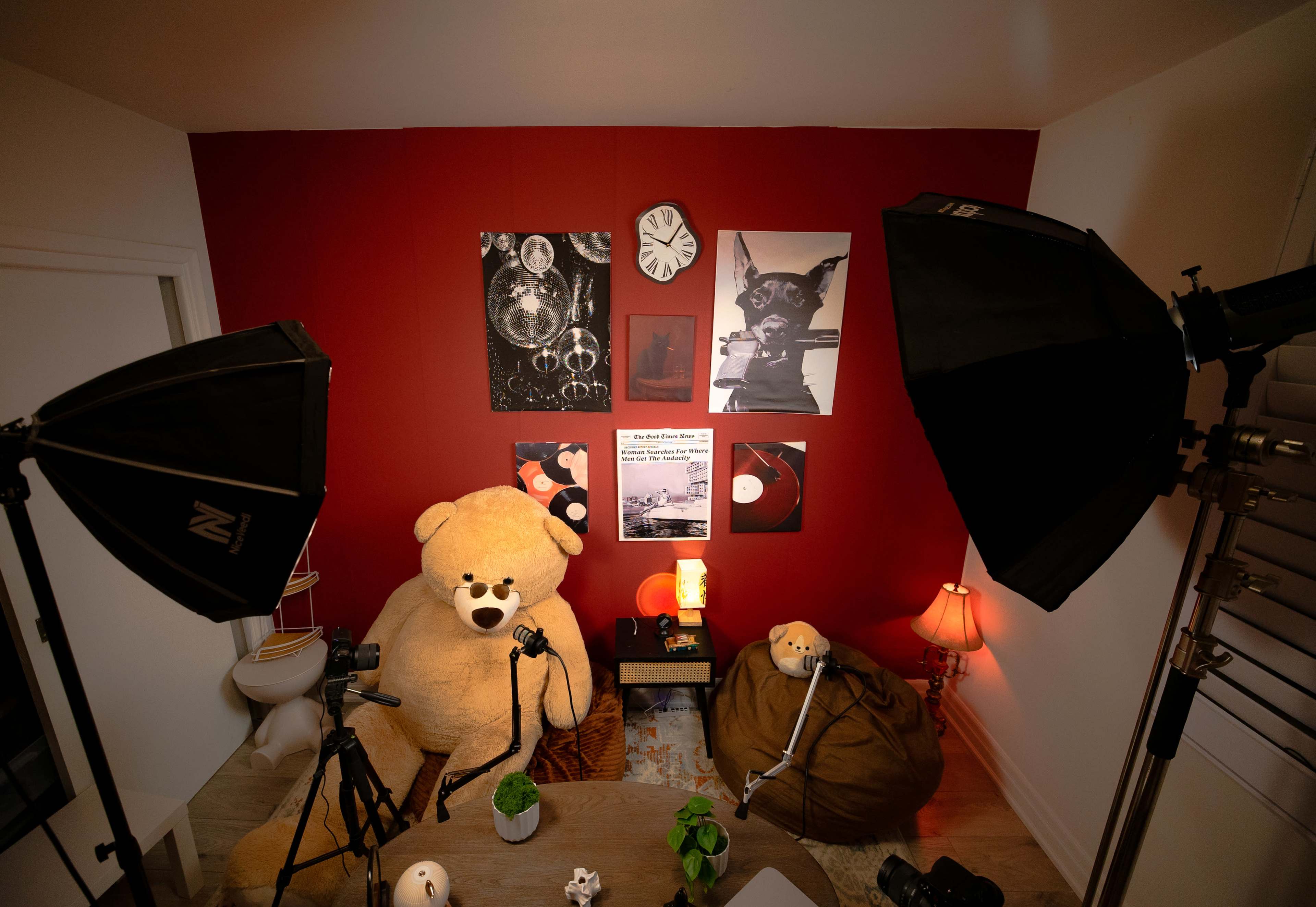 The image shows a cozy room with a large teddy bear, a small table with a lamp, and photo frames on the red wall, illuminated by soft lighting from two studio lights.