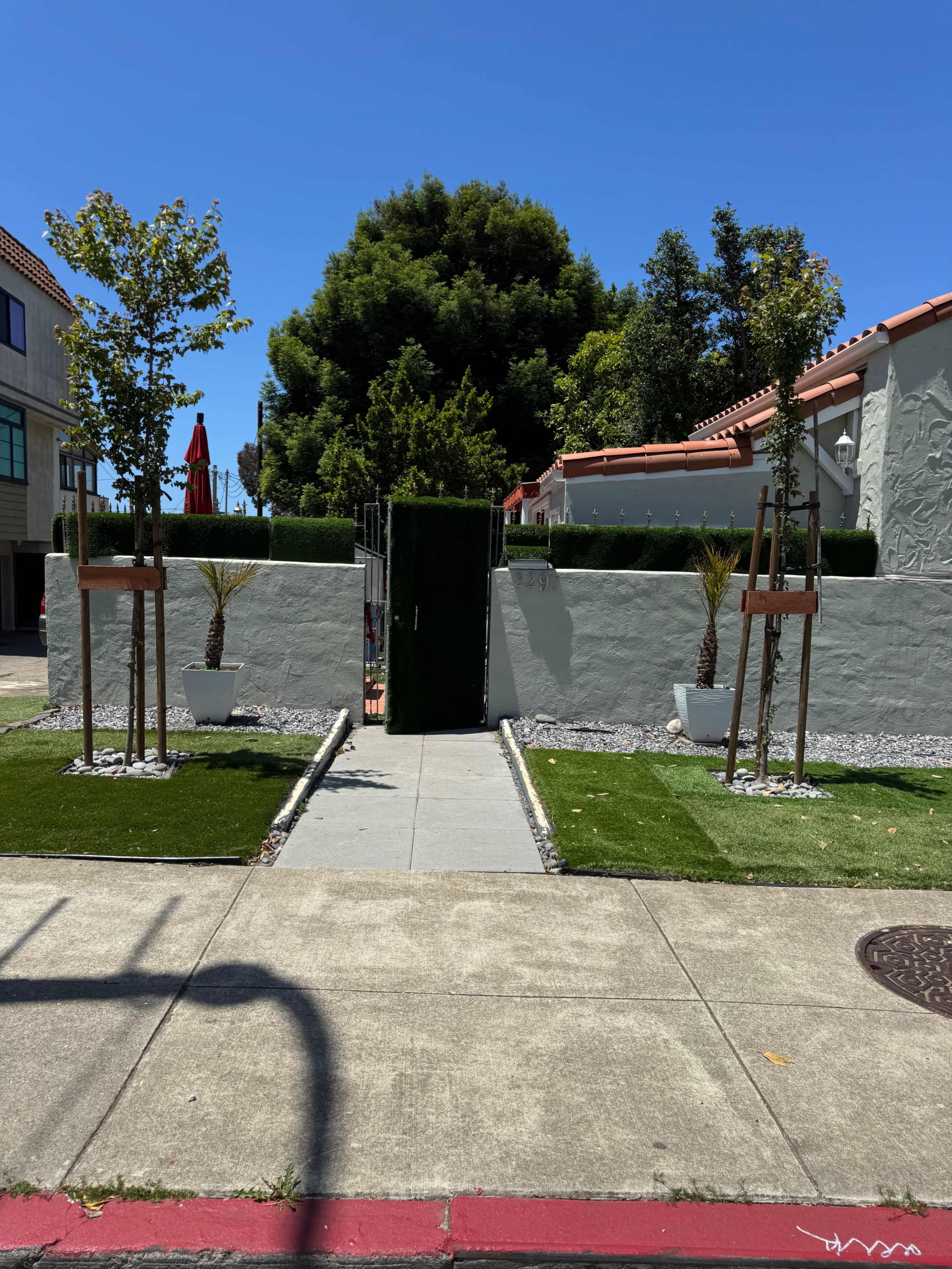 The image shows a neatly arranged entrance pathway flanked by greenery and decorative plants leading to a gated entrance.