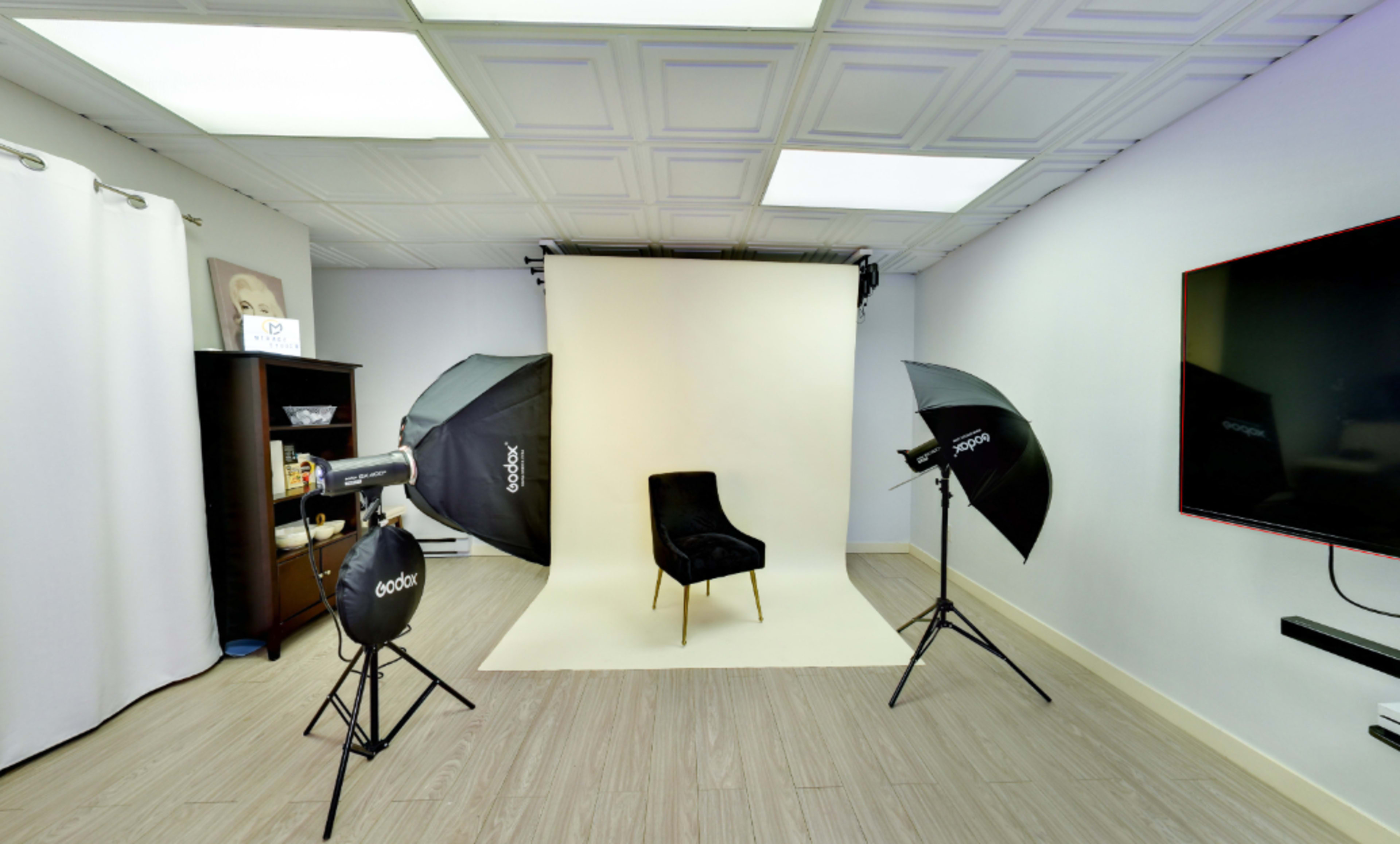 A photographer's studio setup featuring a black chair against a light backdrop, with two softbox lights positioned on either side and a TV mounted on the wall.