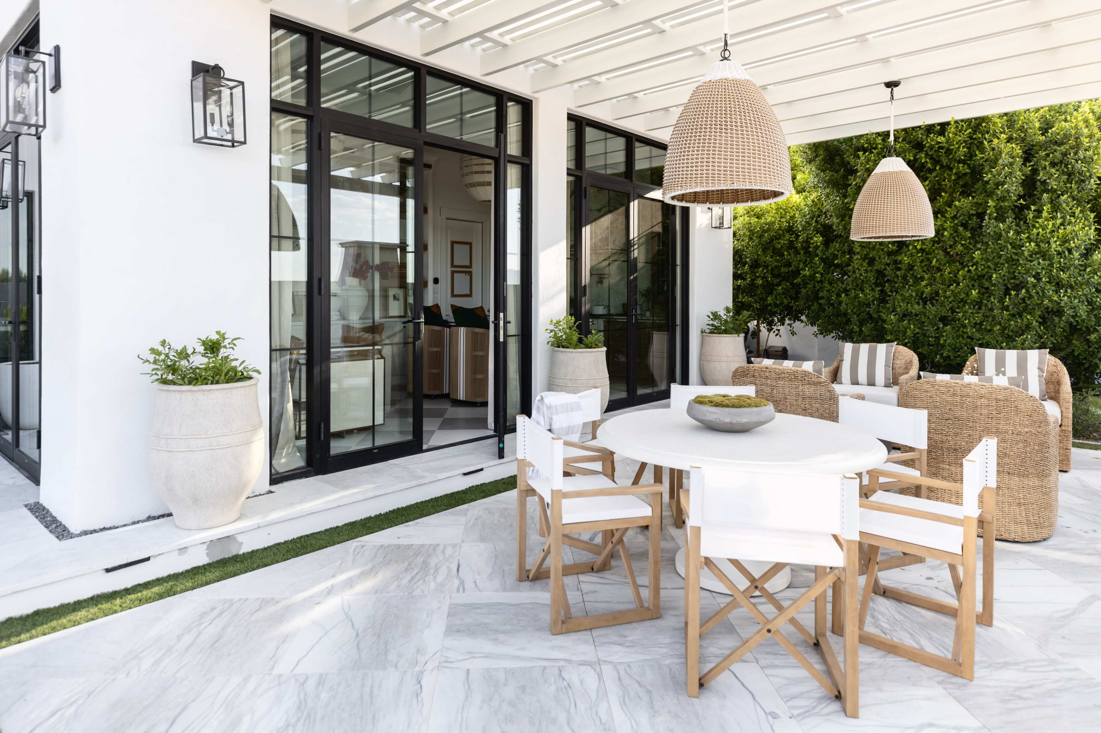 A modern outdoor patio features a round dining table surrounded by white and wooden chairs, with rattan chairs and large planters set against a backdrop of glass doors.