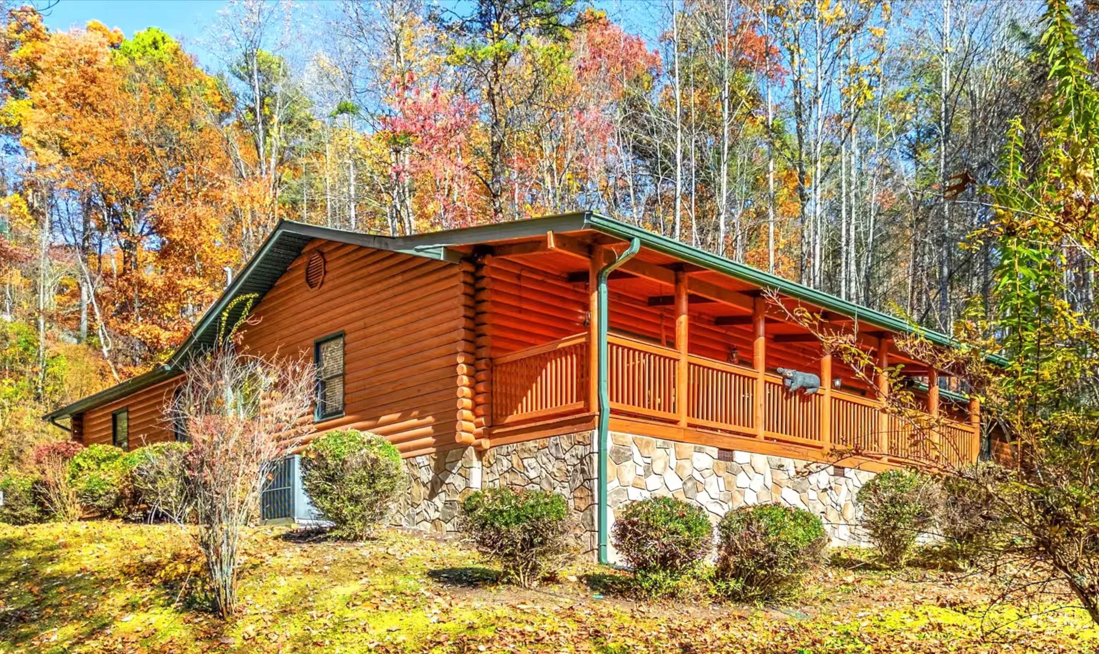 The image shows a log cabin with a stone foundation, situated among trees displaying autumn foliage.