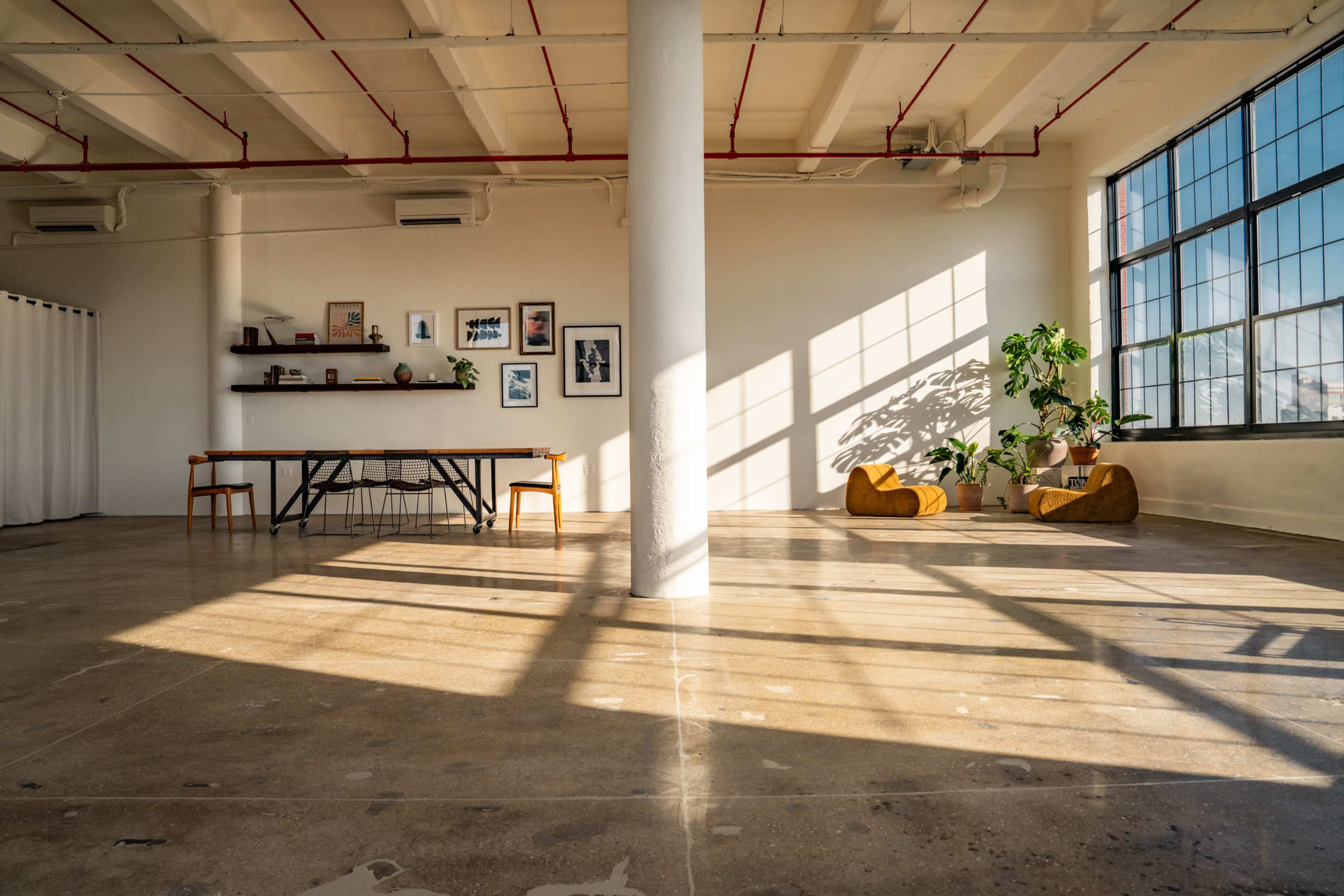 The image shows a spacious, well-lit room with a concrete floor, a long table with chairs, and large windows casting shadows across the space, along with decorative plants and wall art.