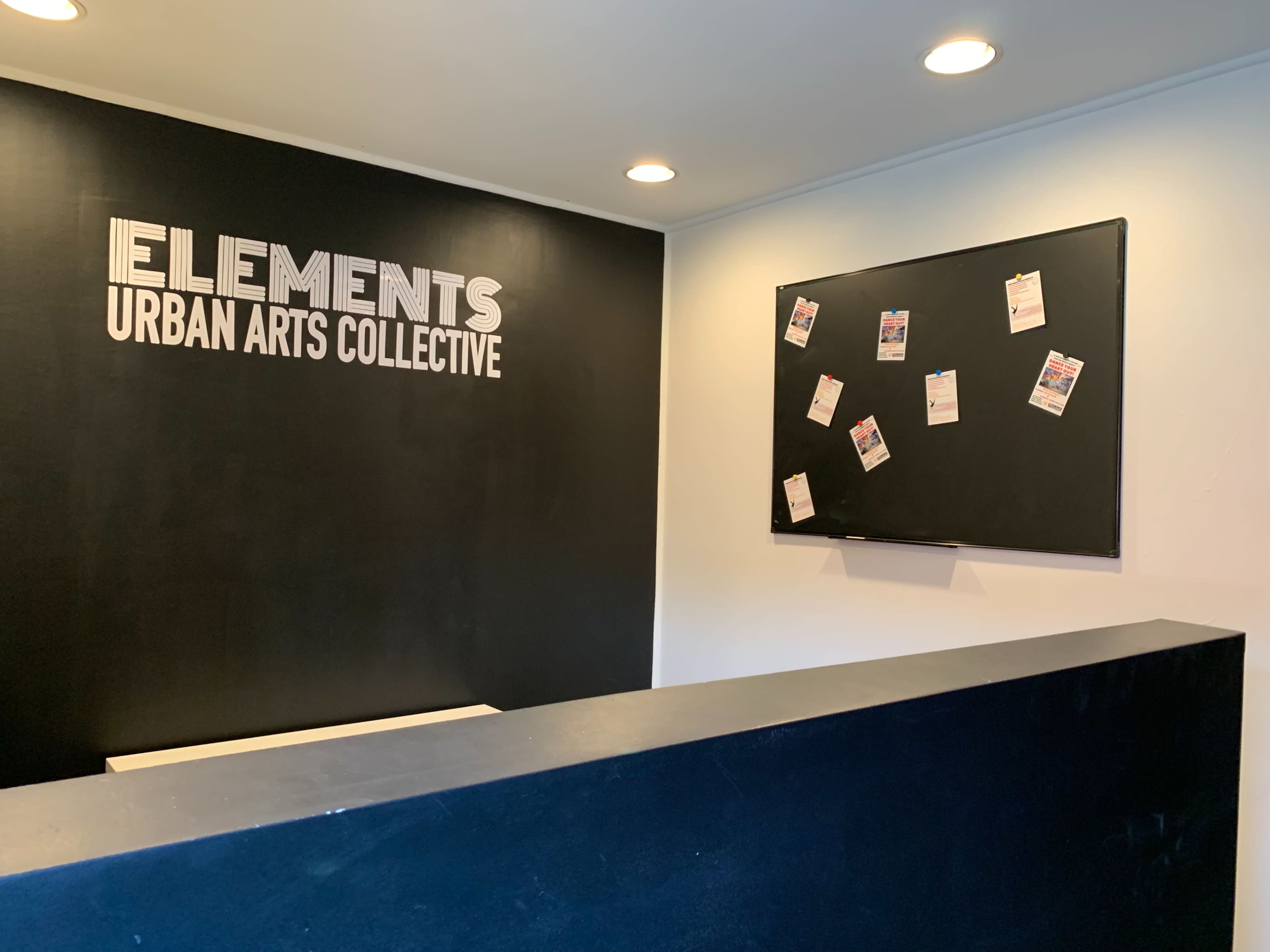 A modern reception area with a black wall displaying the words "ELEMENTS URBAN ARTS COLLECTIVE" and a bulletin board featuring several pinned announcements.