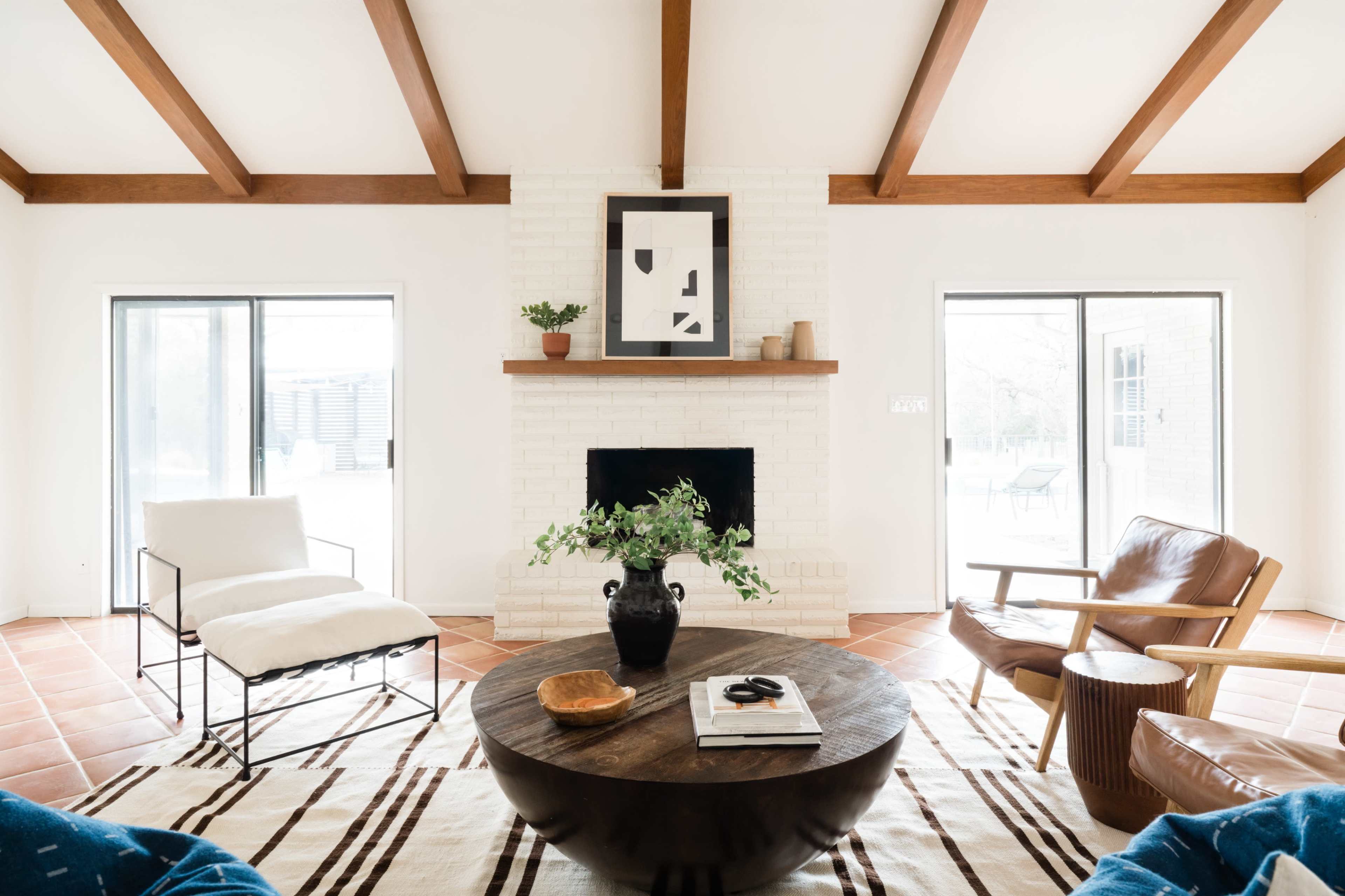 A spacious living room features a coffee table, a white armchair, a brown leather chair, and a brick fireplace, with large windows on either side allowing natural light to enter.