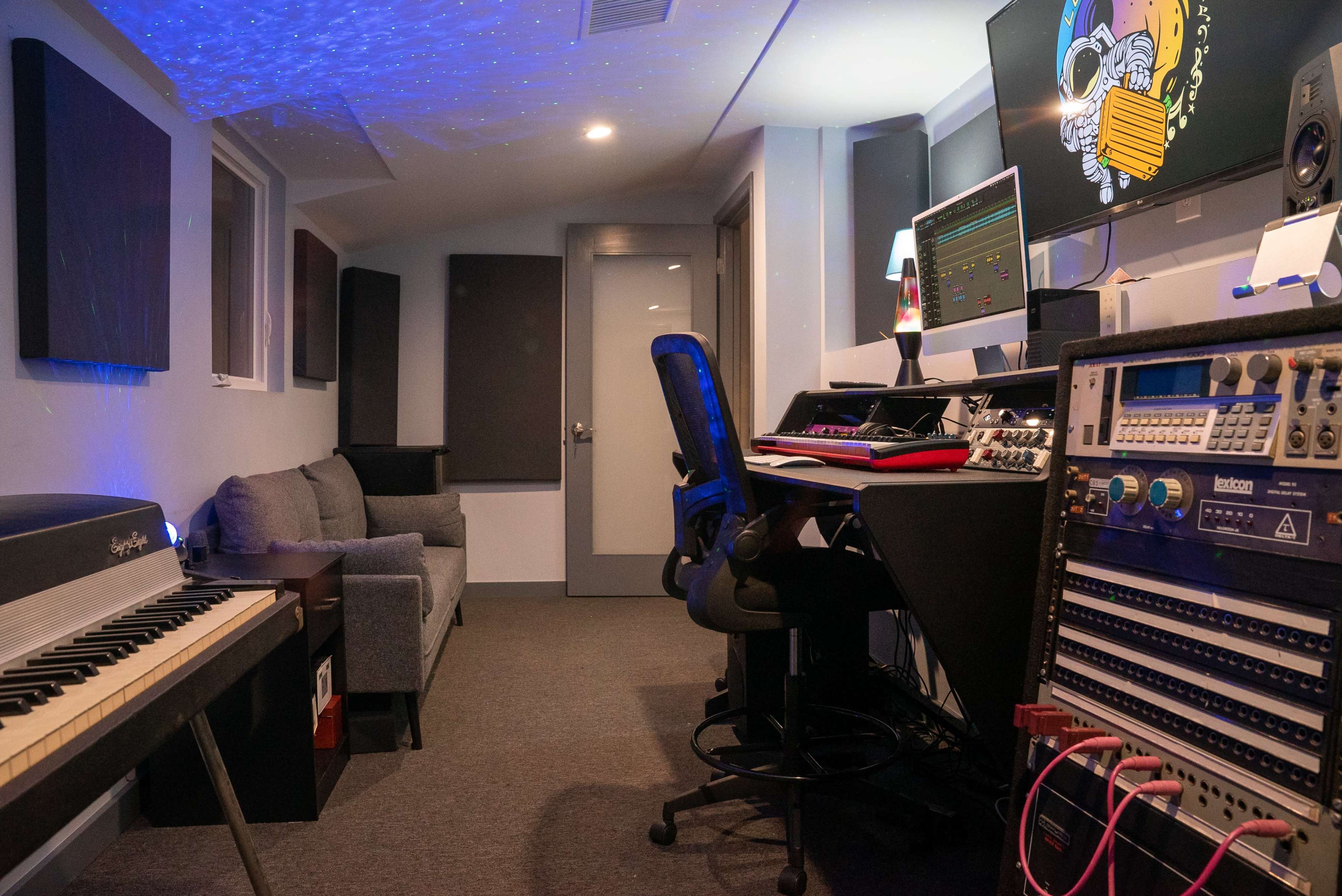 The image shows a recording studio with a keyboard, computer monitors, audio equipment, a black office chair, and a gray sofa against the wall.