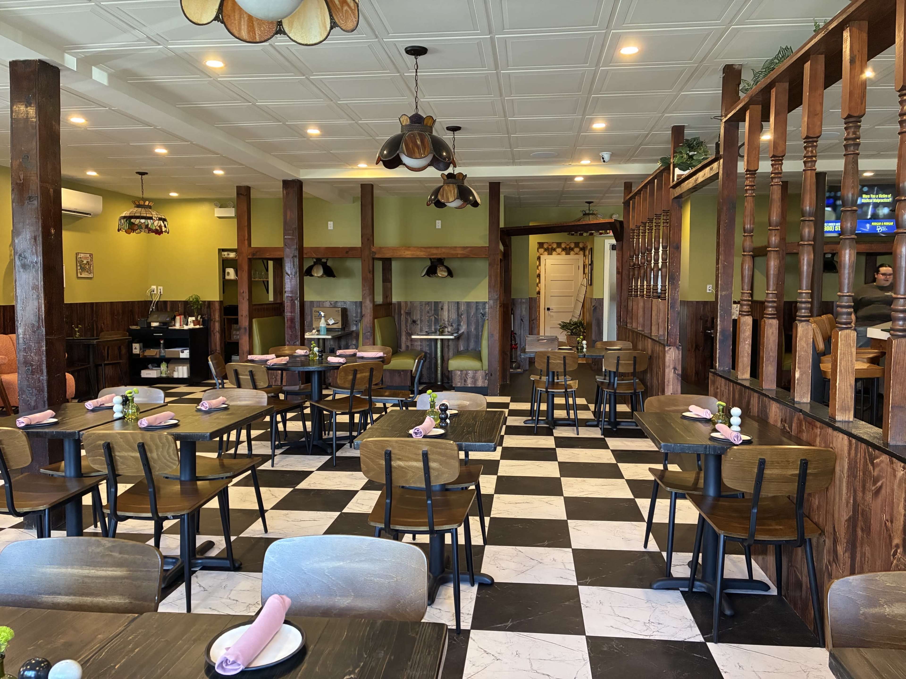 The image shows a restaurant interior featuring a mix of wooden tables and chairs arranged on a black-and-white checkered floor, with green walls, decorative lighting, and a seating area in the background.