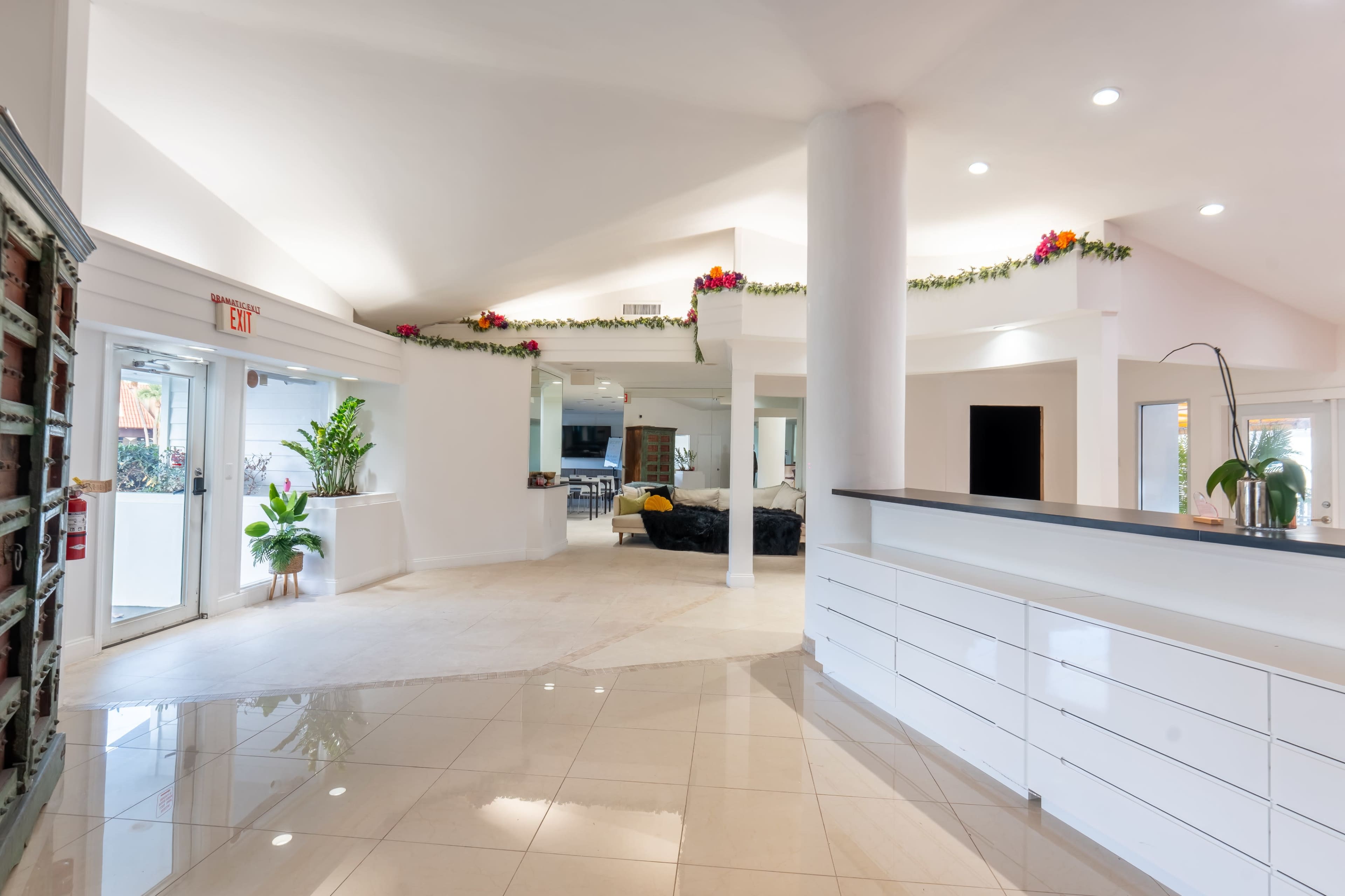 The image shows a spacious, brightly lit lobby area with white walls, a tiled floor, and decorative plants.