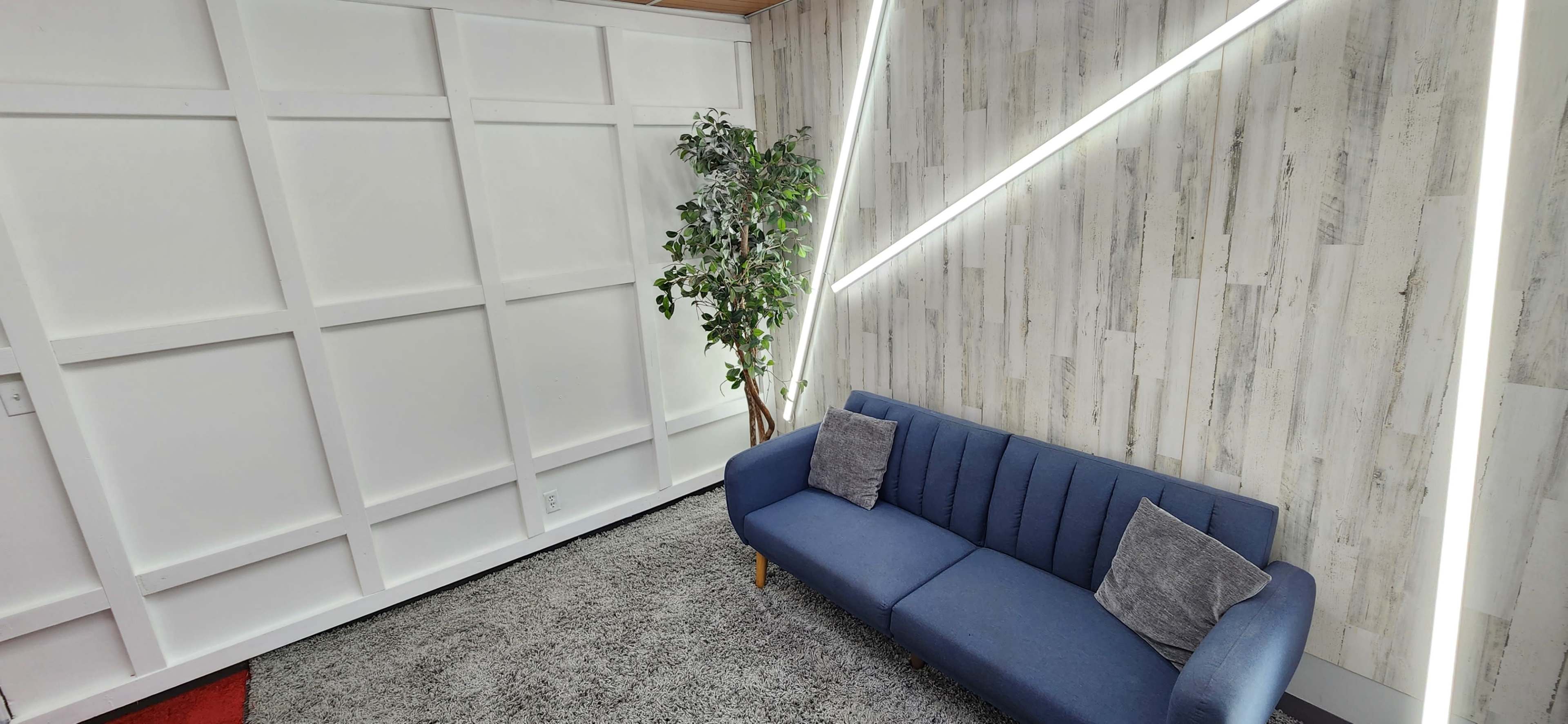 The image shows a modern lounge area featuring a blue couch with two gray pillows, a potted plant, and a wall decorated with white paneling and angled LED lights.