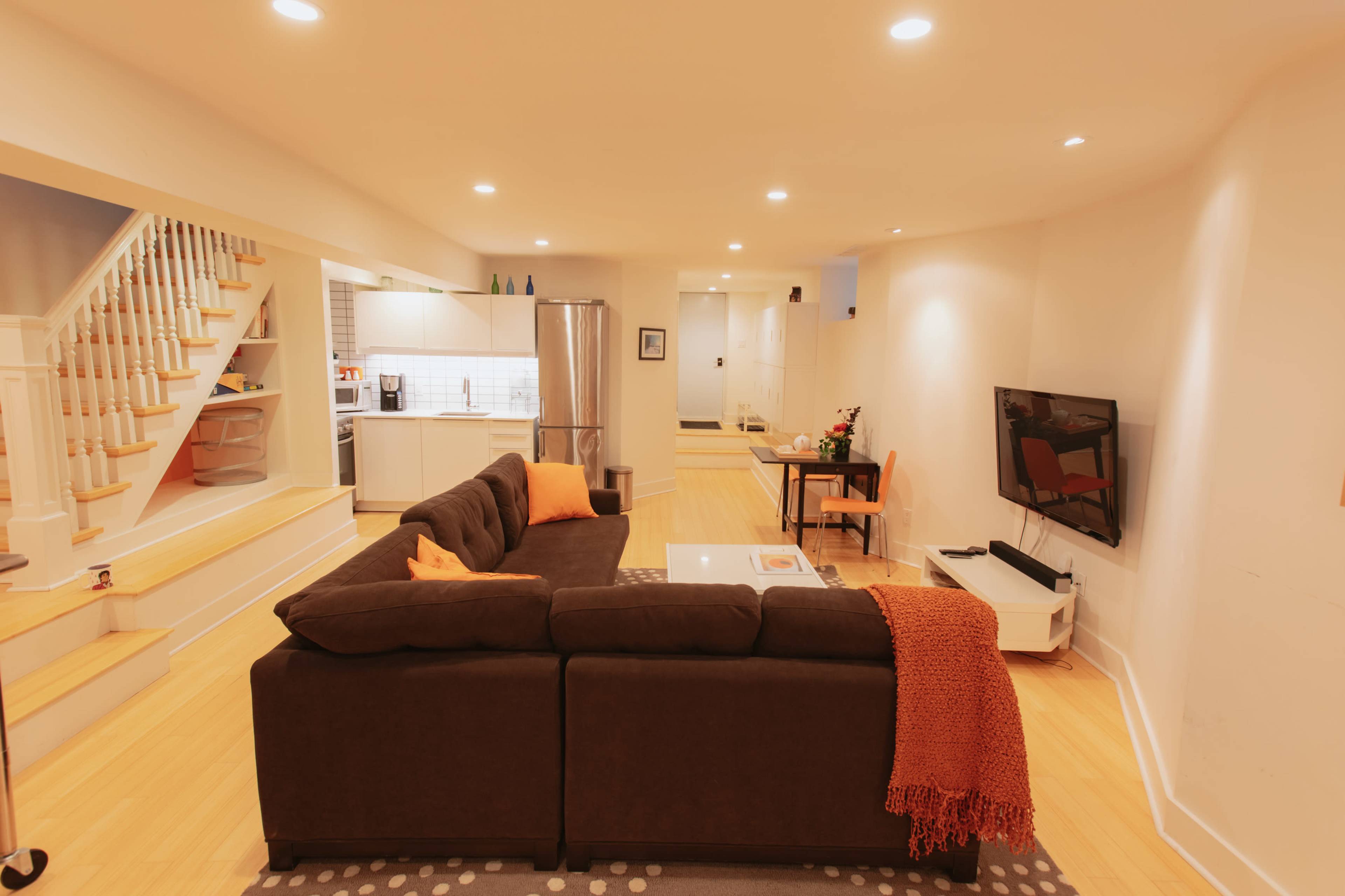 A modern living room features a dark sectional sofa with orange accents, a coffee table, and a television, with a staircase and kitchen visible in the background.