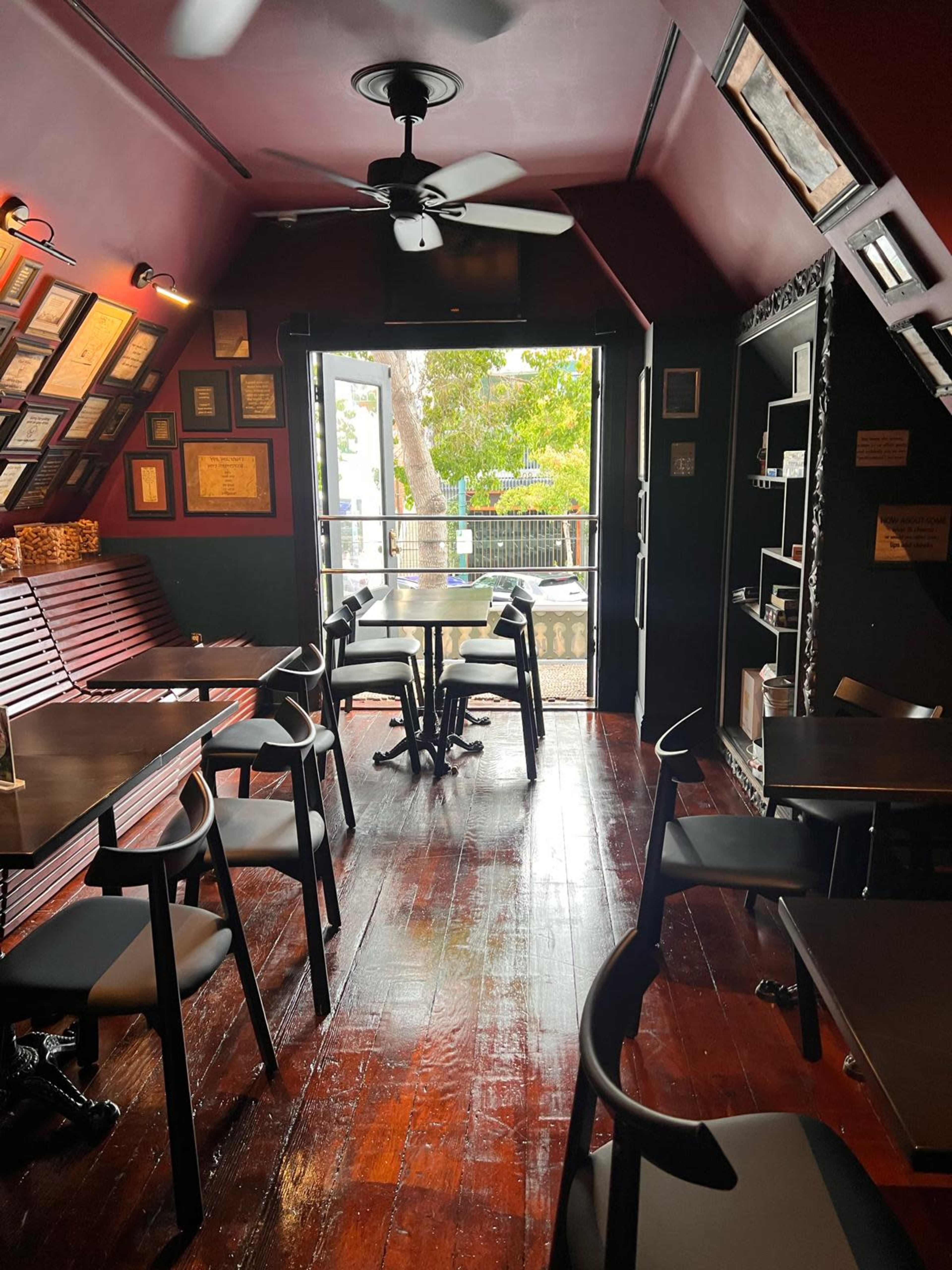 The image shows a cozy dining area with wooden floors, dark walls adorned with certificates, and an open door leading to an outdoor view.