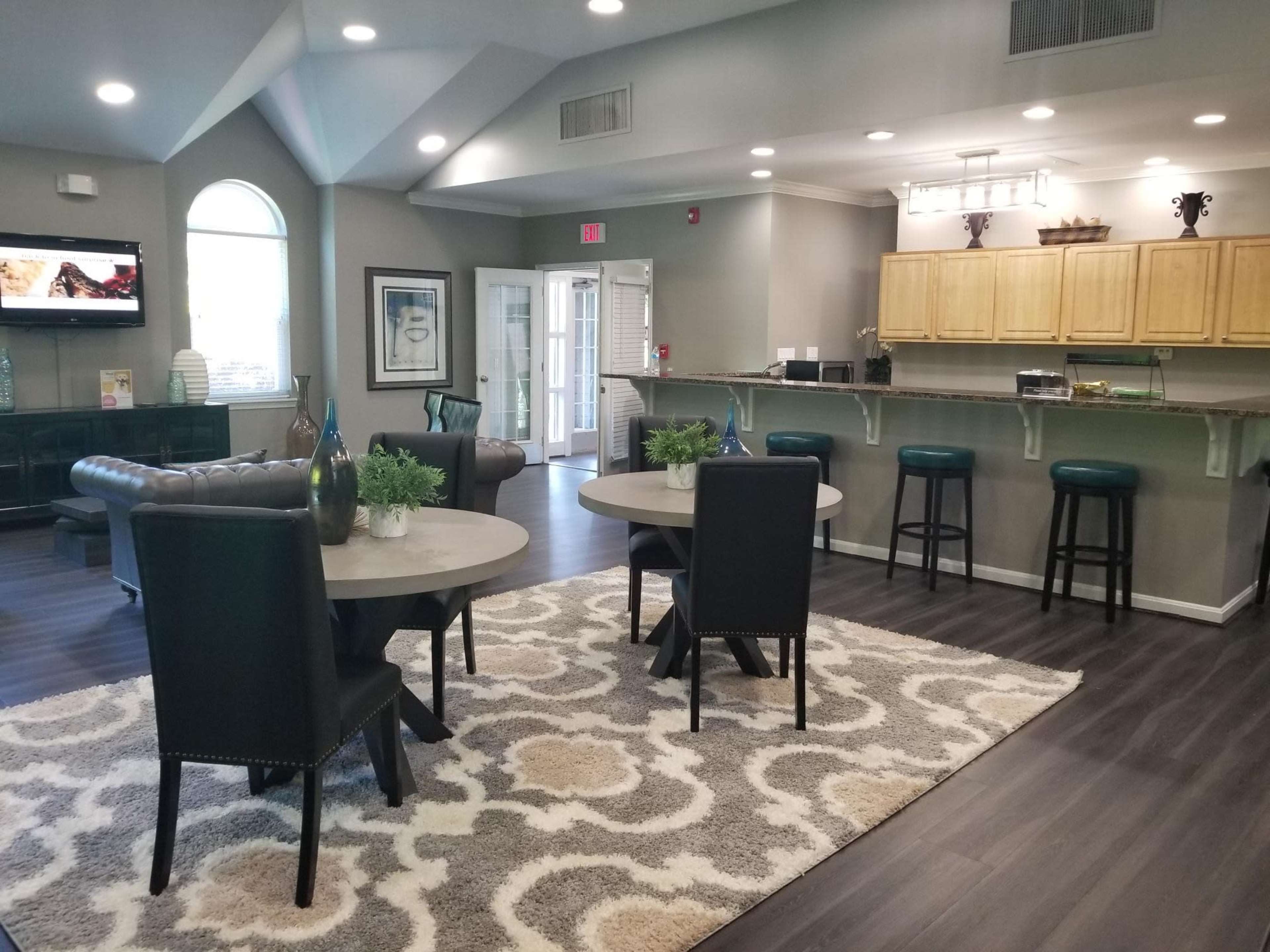A modern, brightly lit communal area with two tables, seating, a rug, and a kitchenette.