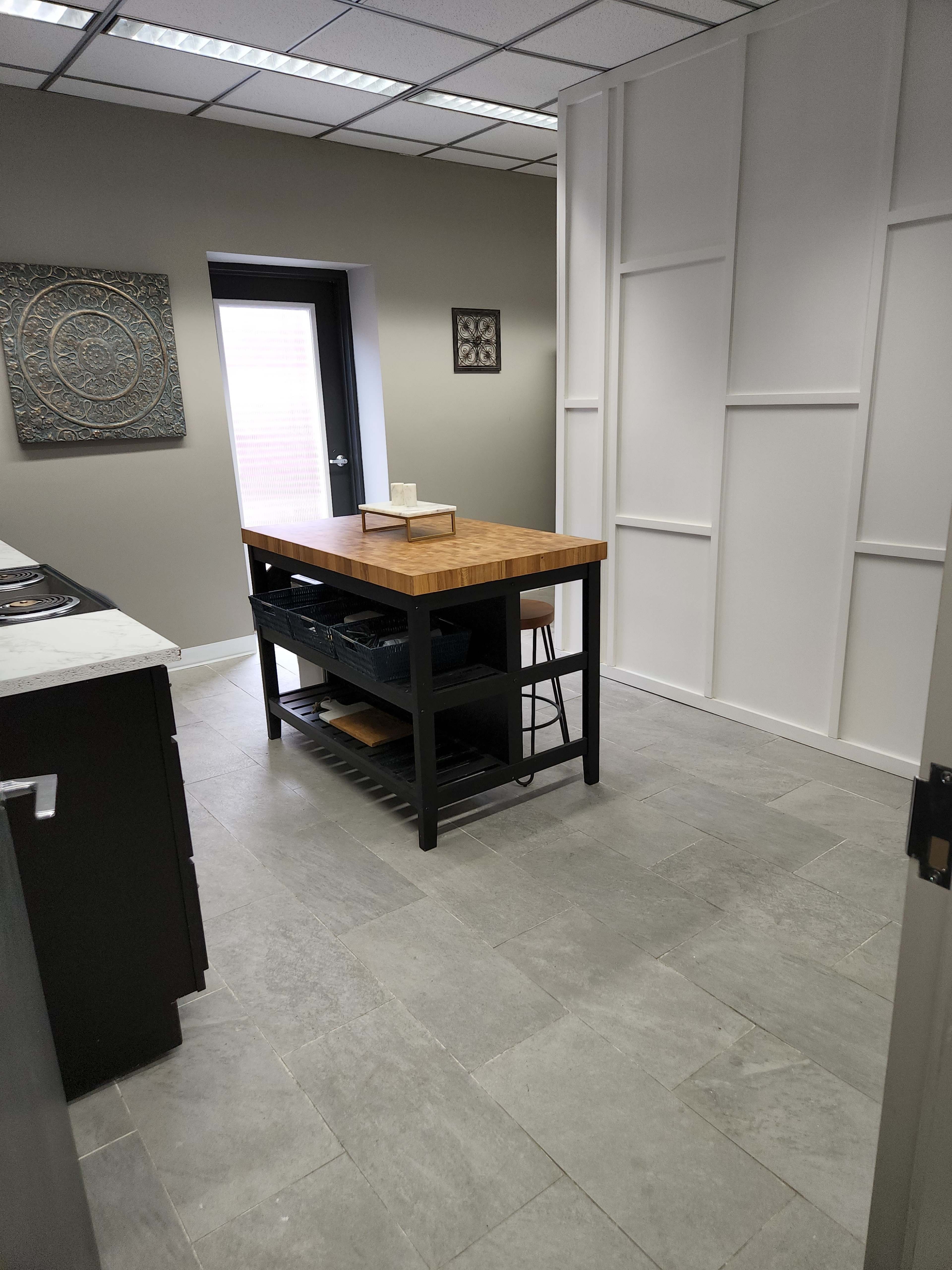 The image shows a modern kitchen with a central wooden island, gray tiled floors, and minimalist decor on the walls.
