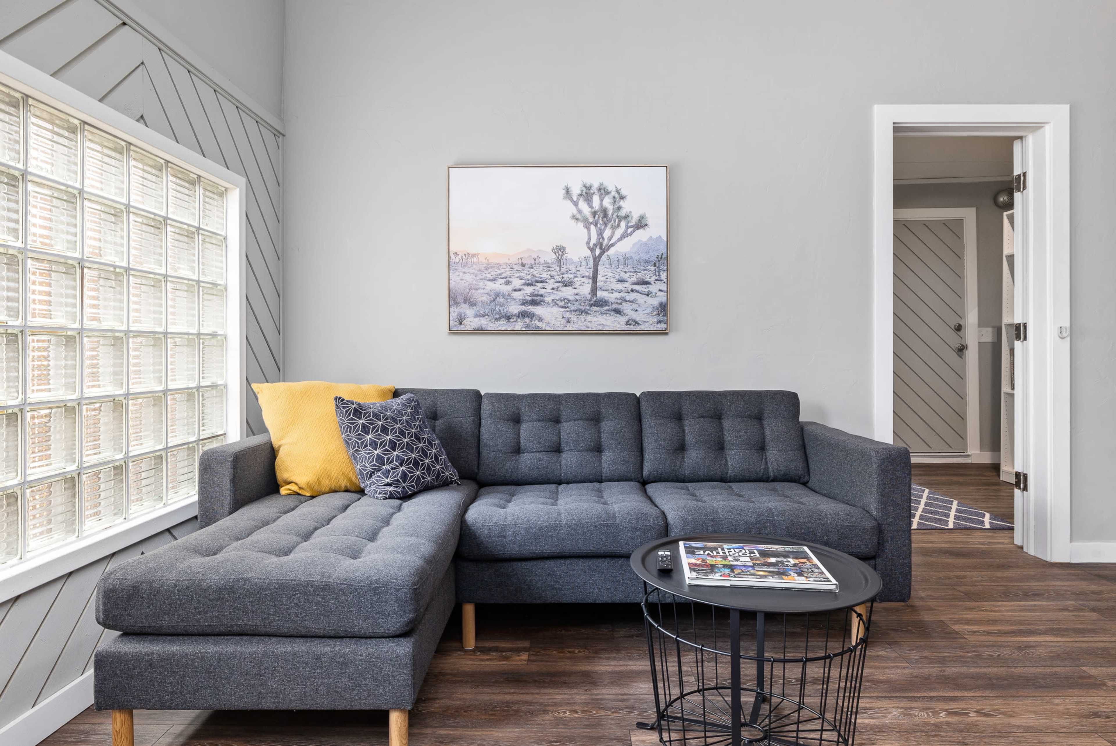 The image shows a modern living room featuring a gray sectional sofa, a small black round table with magazines, and a large window with glass blocks.
