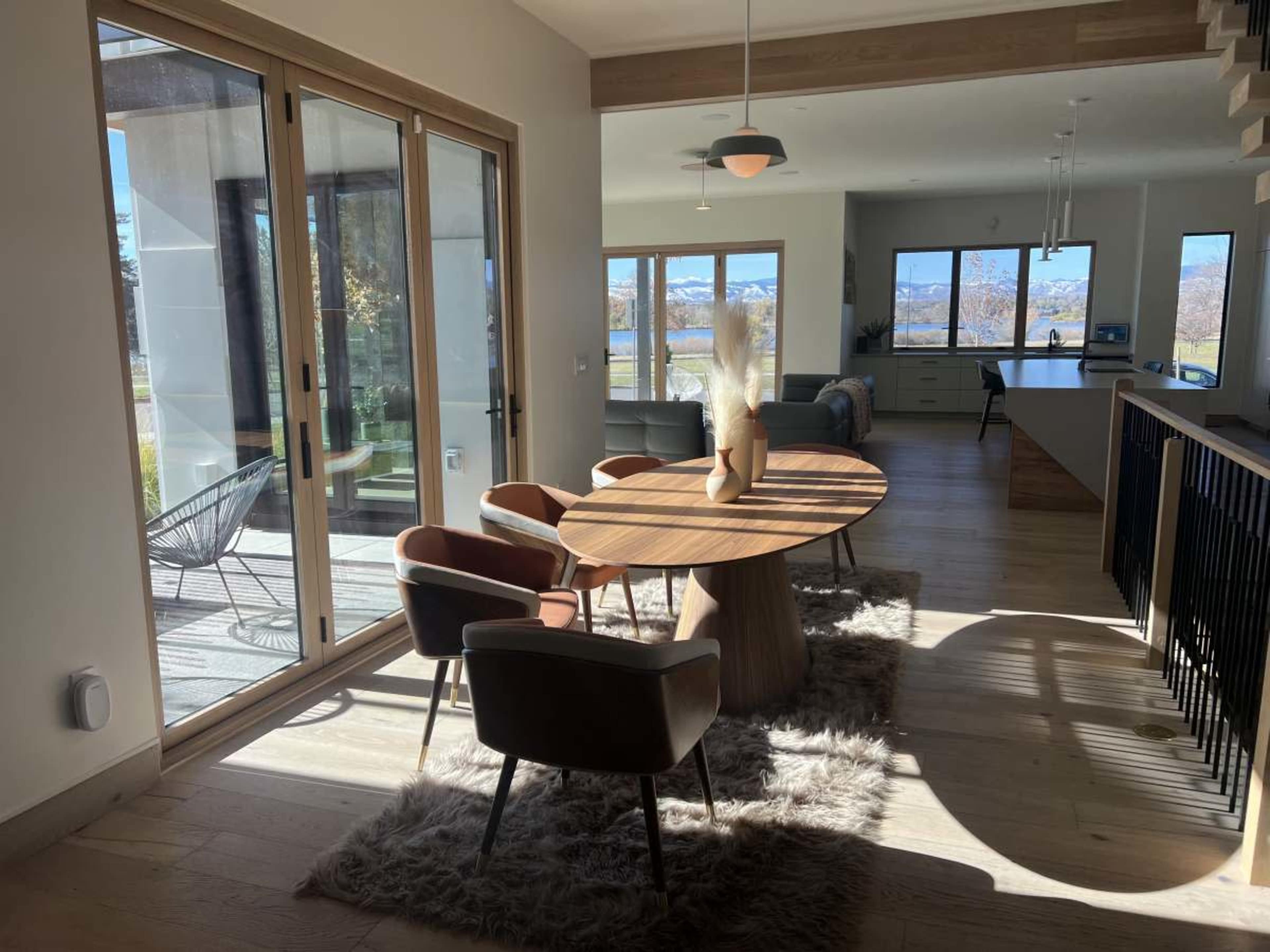 The image showcases a modern interior featuring a circular wooden dining table surrounded by upholstered chairs, with large glass doors leading to an outdoor area and mountains visible in the background.