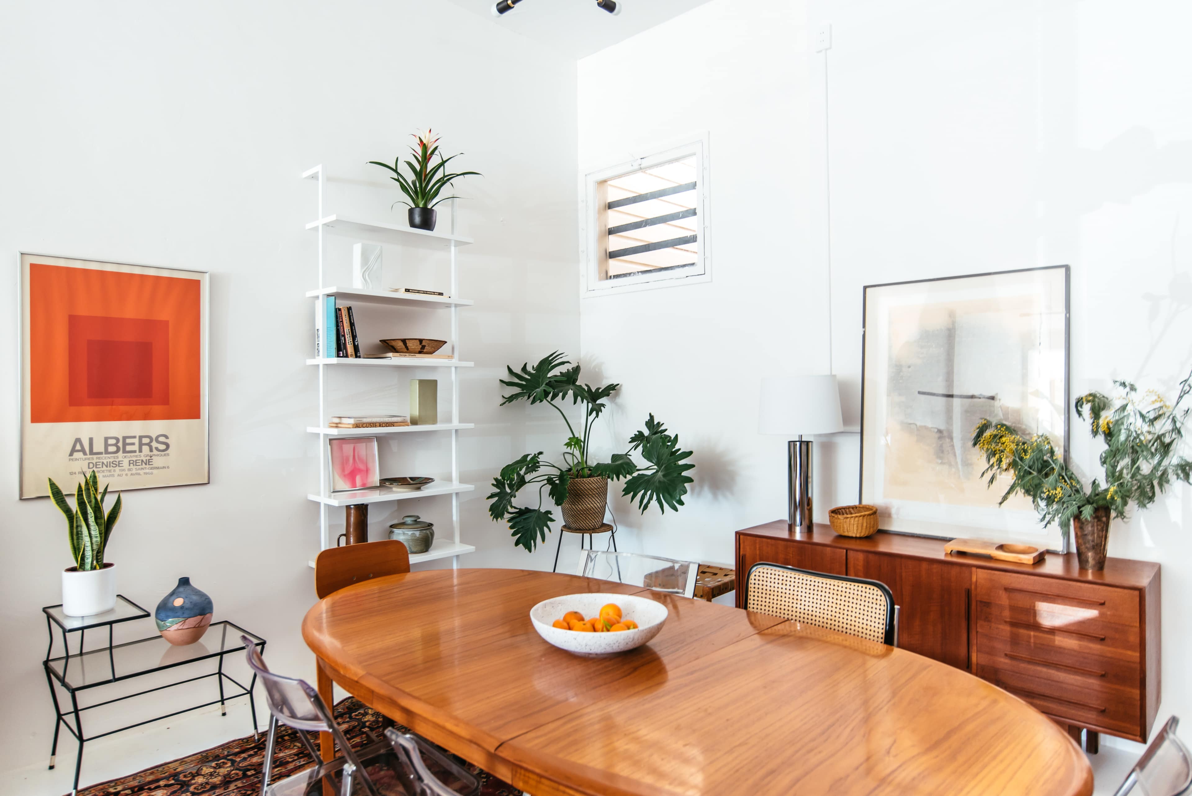 A modern dining area featuring a round wooden table, chairs, and a minimalist shelf with plants and decor against a white wall.