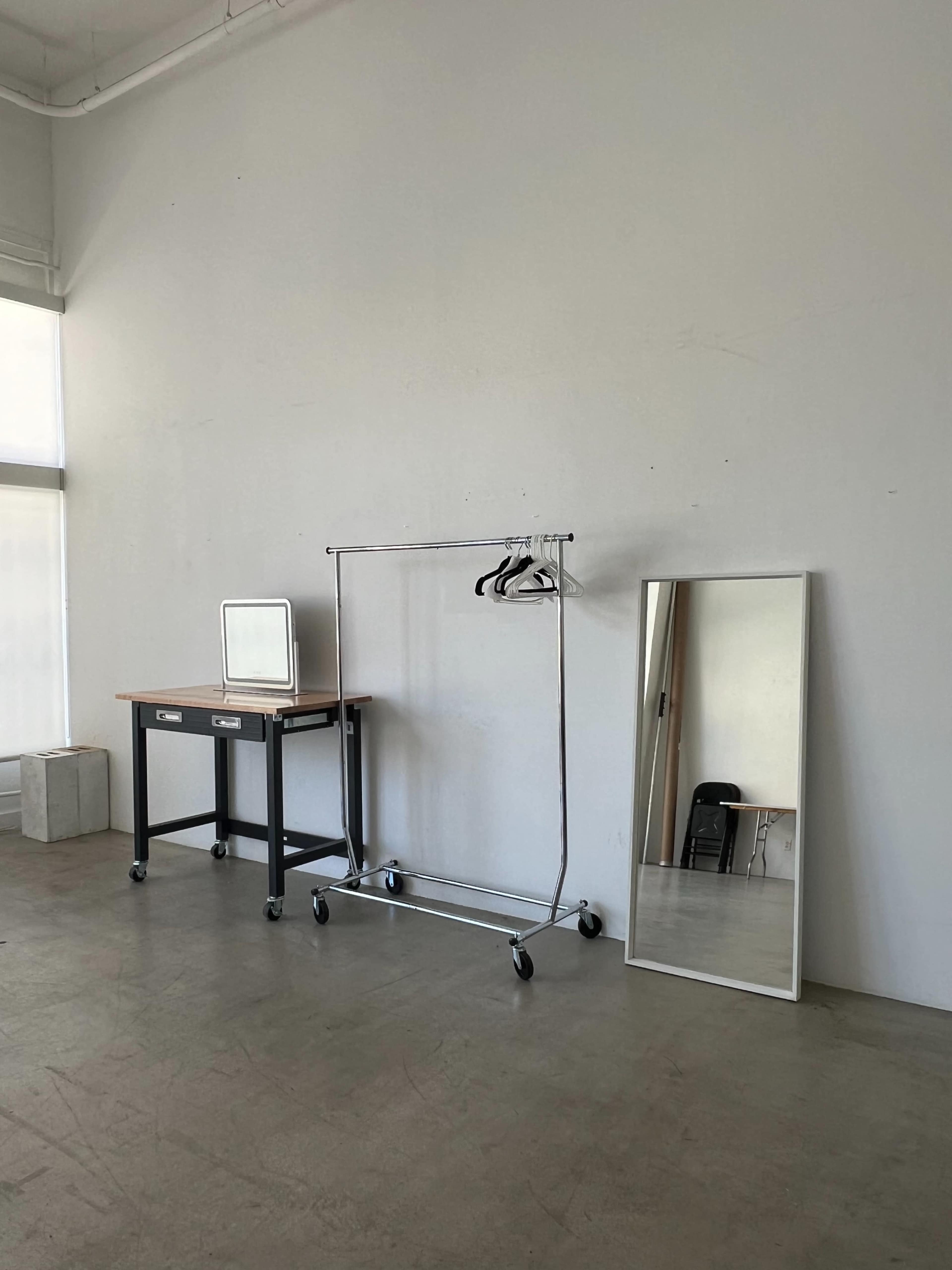 A minimalist room with a mirror, a clothing rack on wheels with three hangers, and a small table with a mirror against a plain wall.