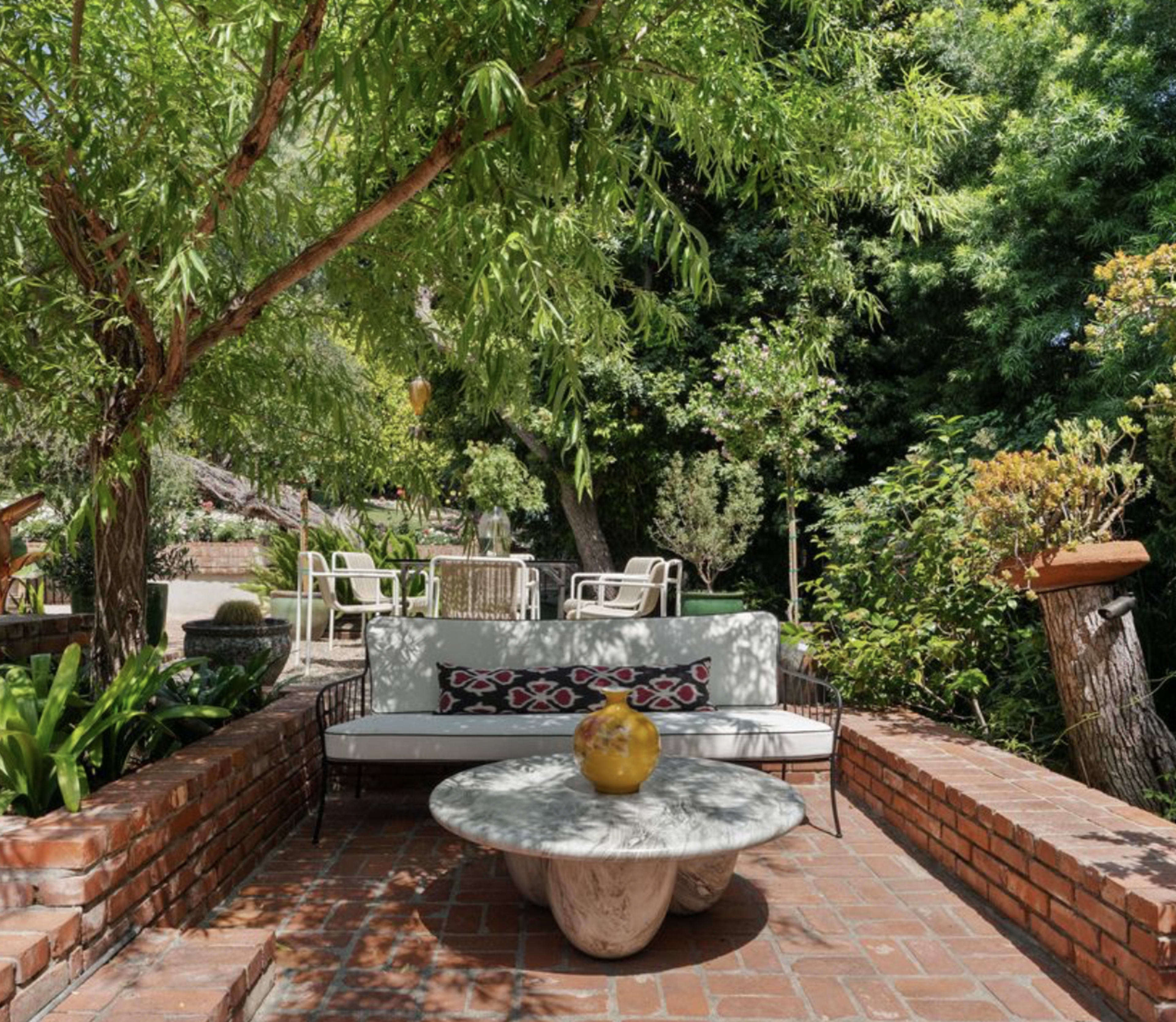 The image shows a brick patio with a seating area, featuring a stone table and a bench, surrounded by lush greenery and trees.