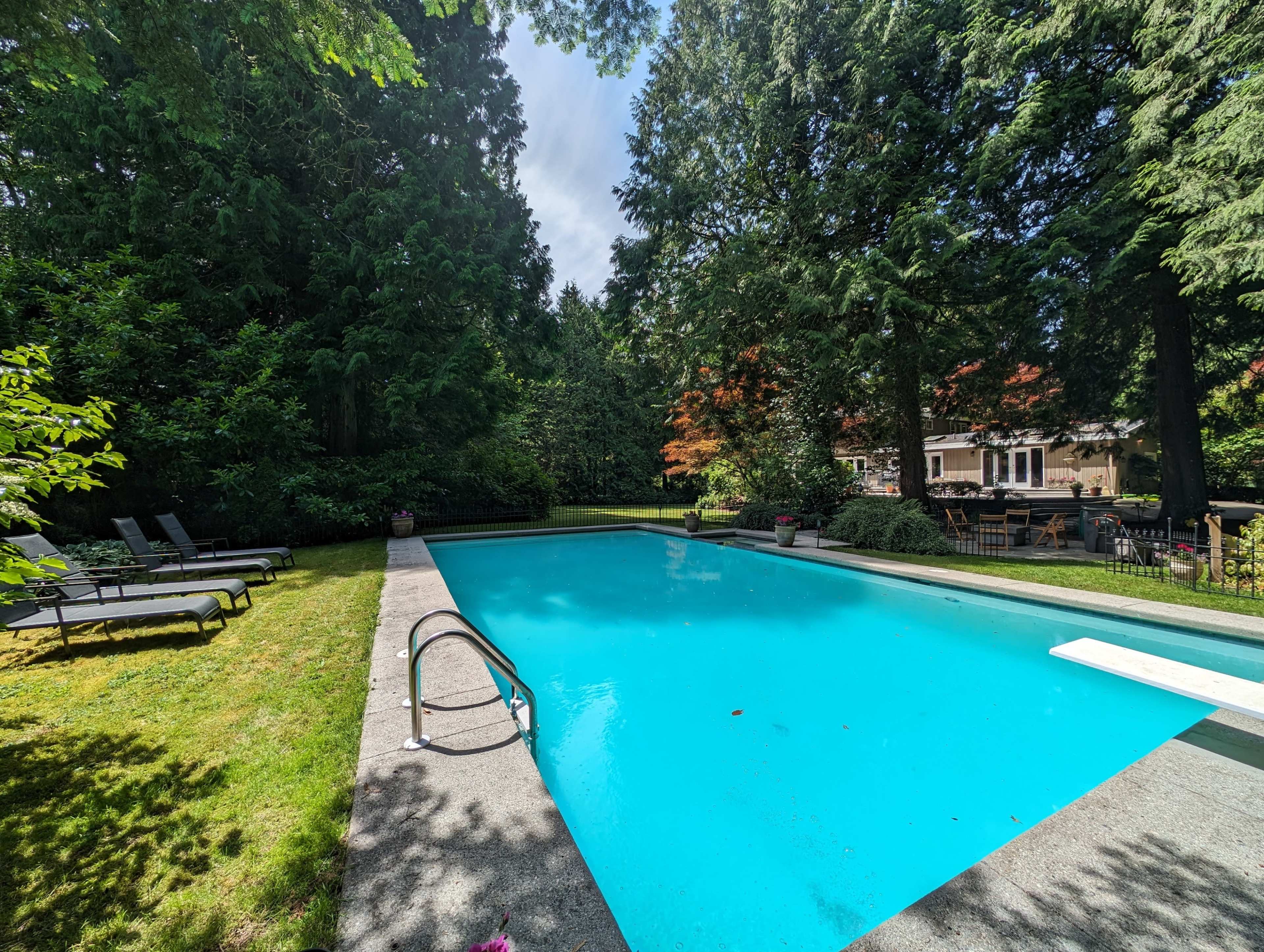 A swimming pool with clear blue water is surrounded by greenery and lawn chairs in a landscaped backyard.