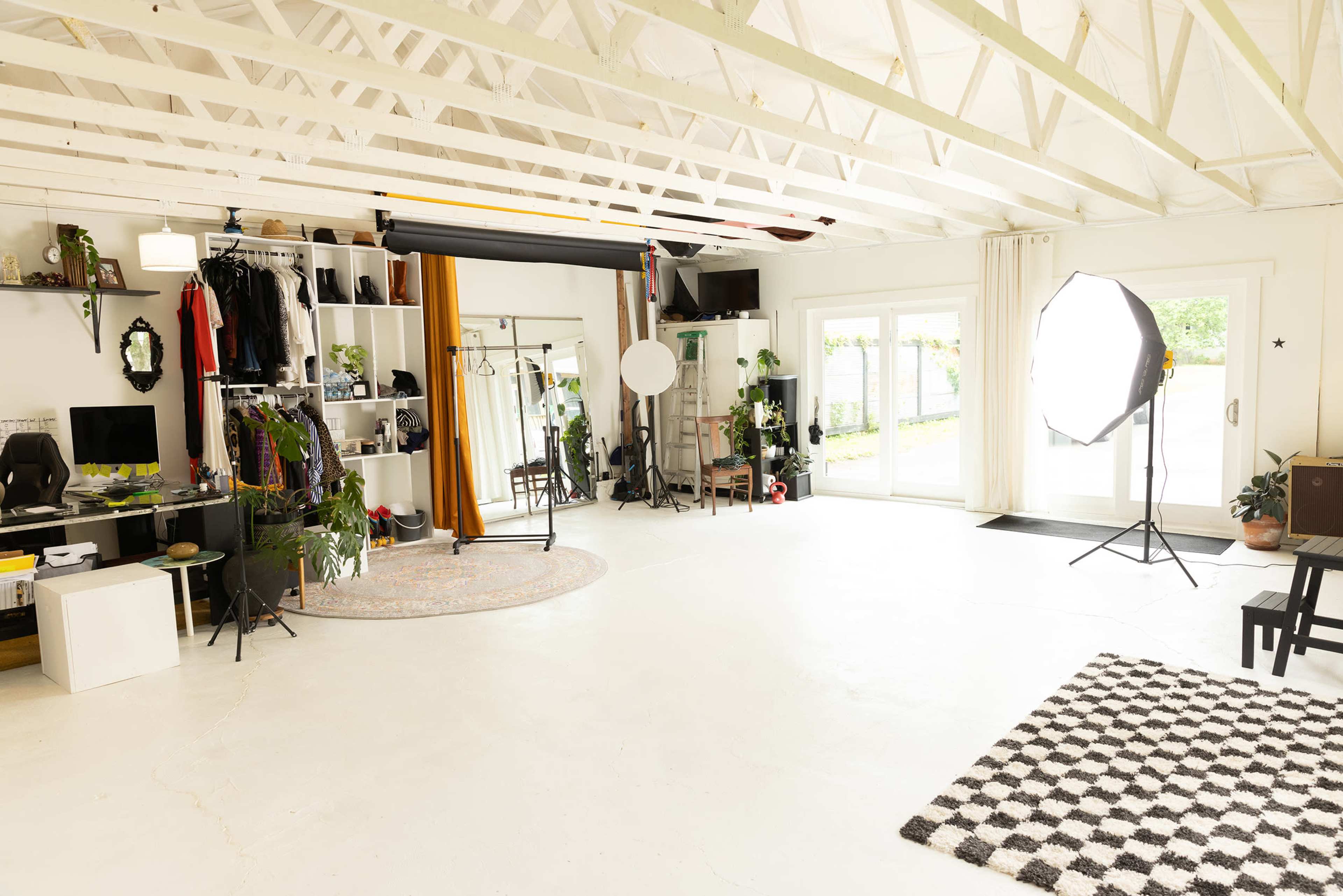 The image shows a spacious, well-lit studio with a variety of clothing, plants, and photography equipment arranged around the room.