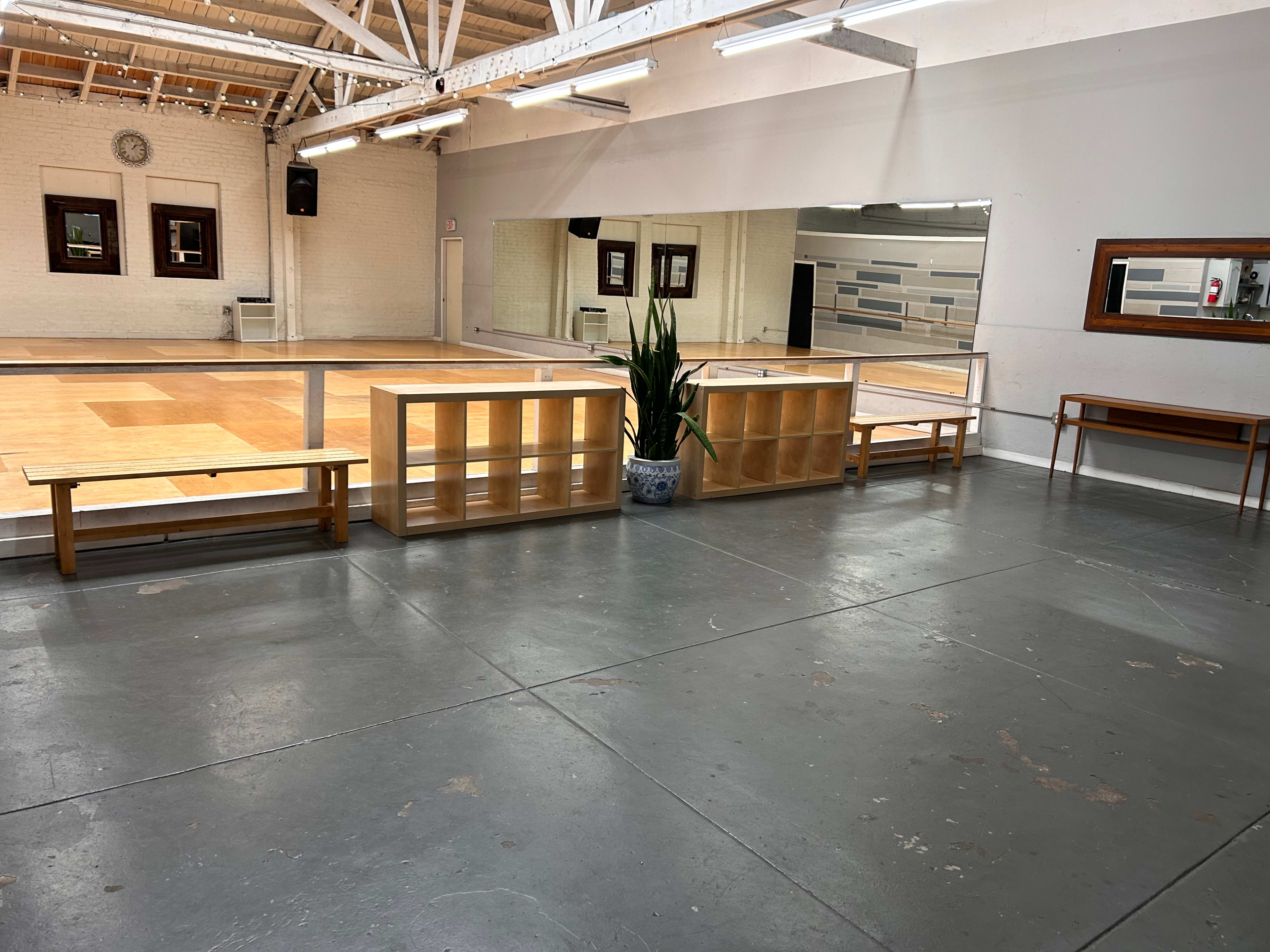 A spacious dance studio features wooden flooring, large mirrors, and benches arranged around potted plants.