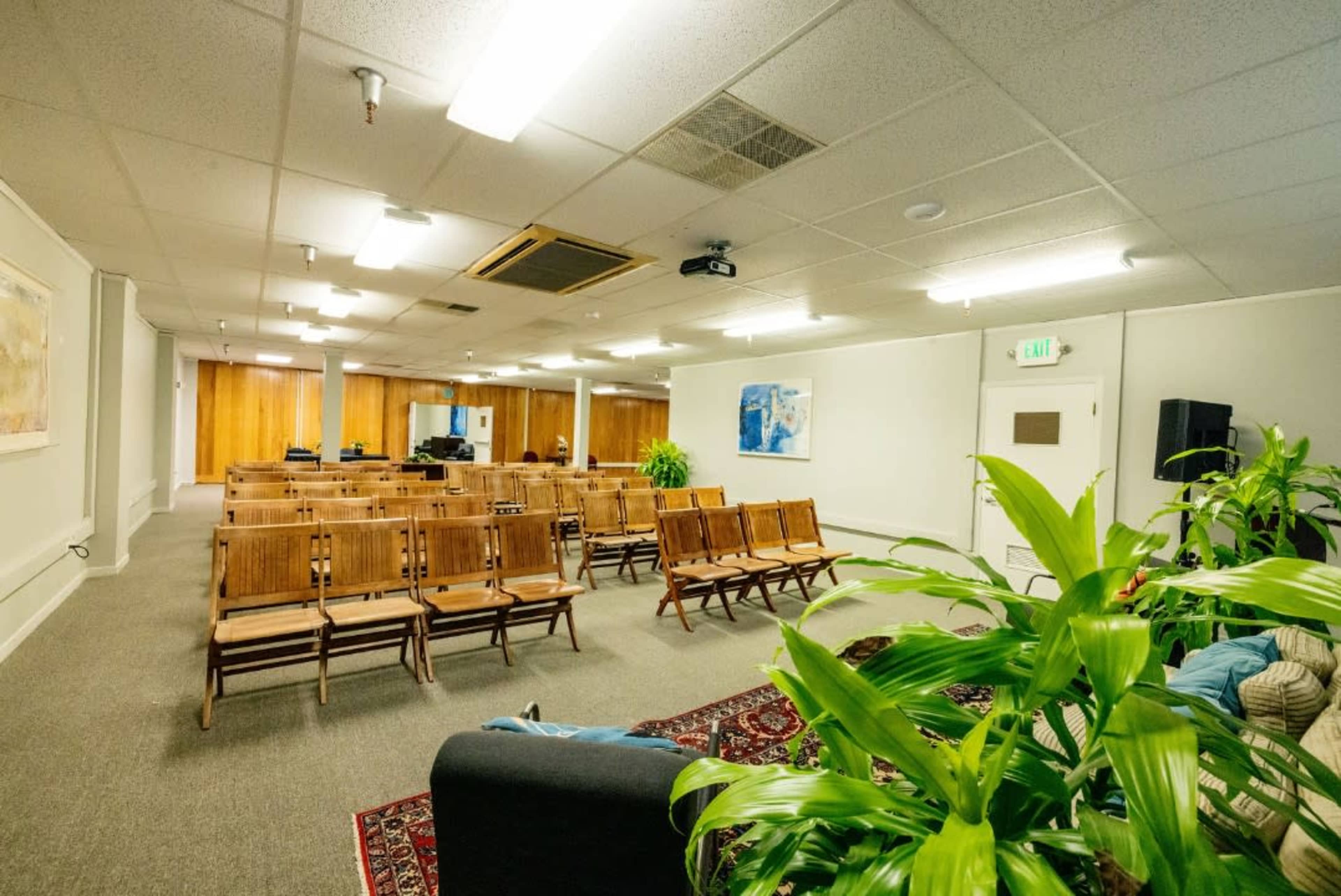 A sparsely furnished meeting room with wooden chairs arranged in rows and potted plants in the corners.