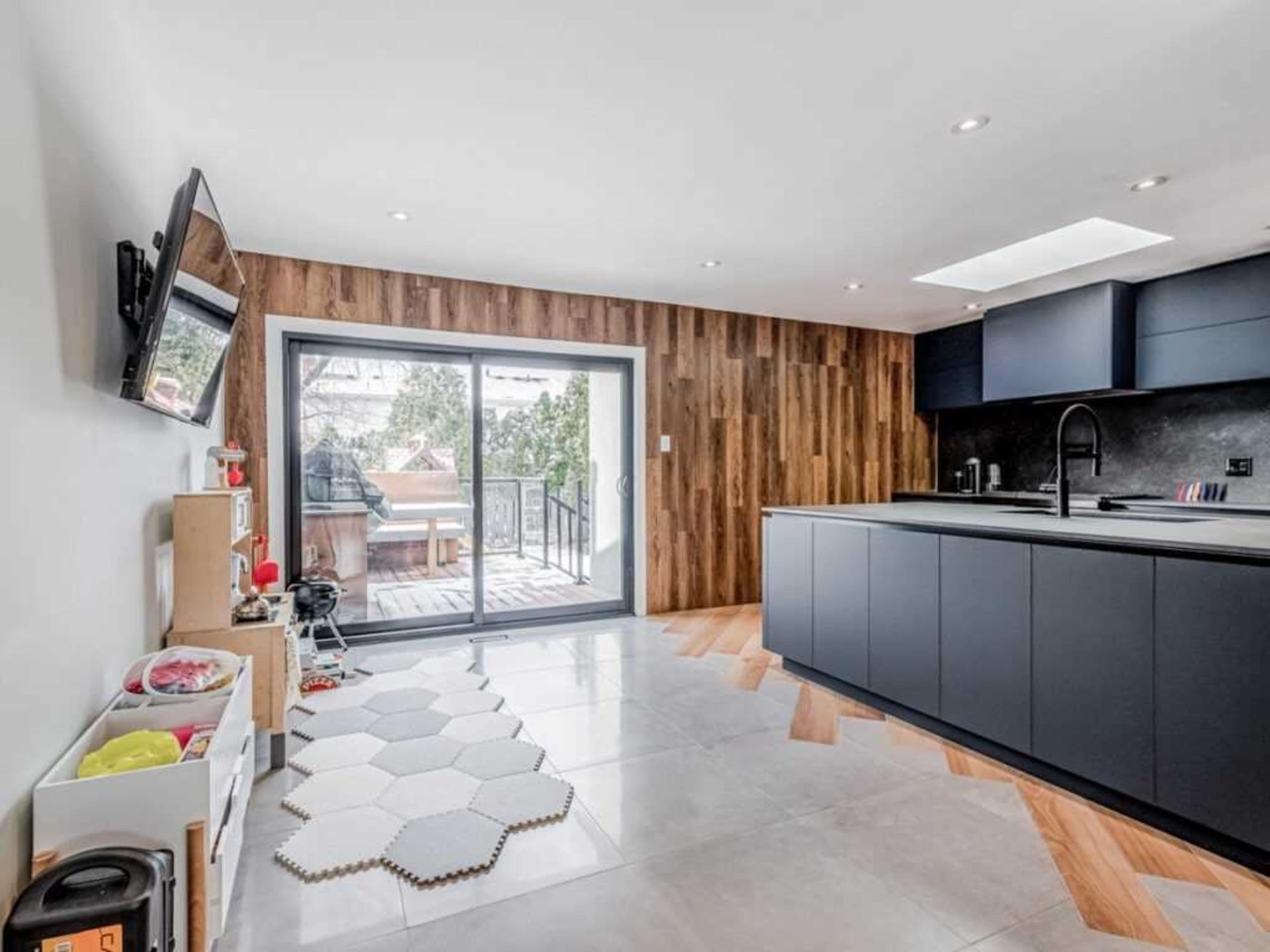 The image shows a modern kitchen featuring a dark cabinetry layout, a sliding glass door leading to a deck, and a light-colored hexagonal rug on the floor.