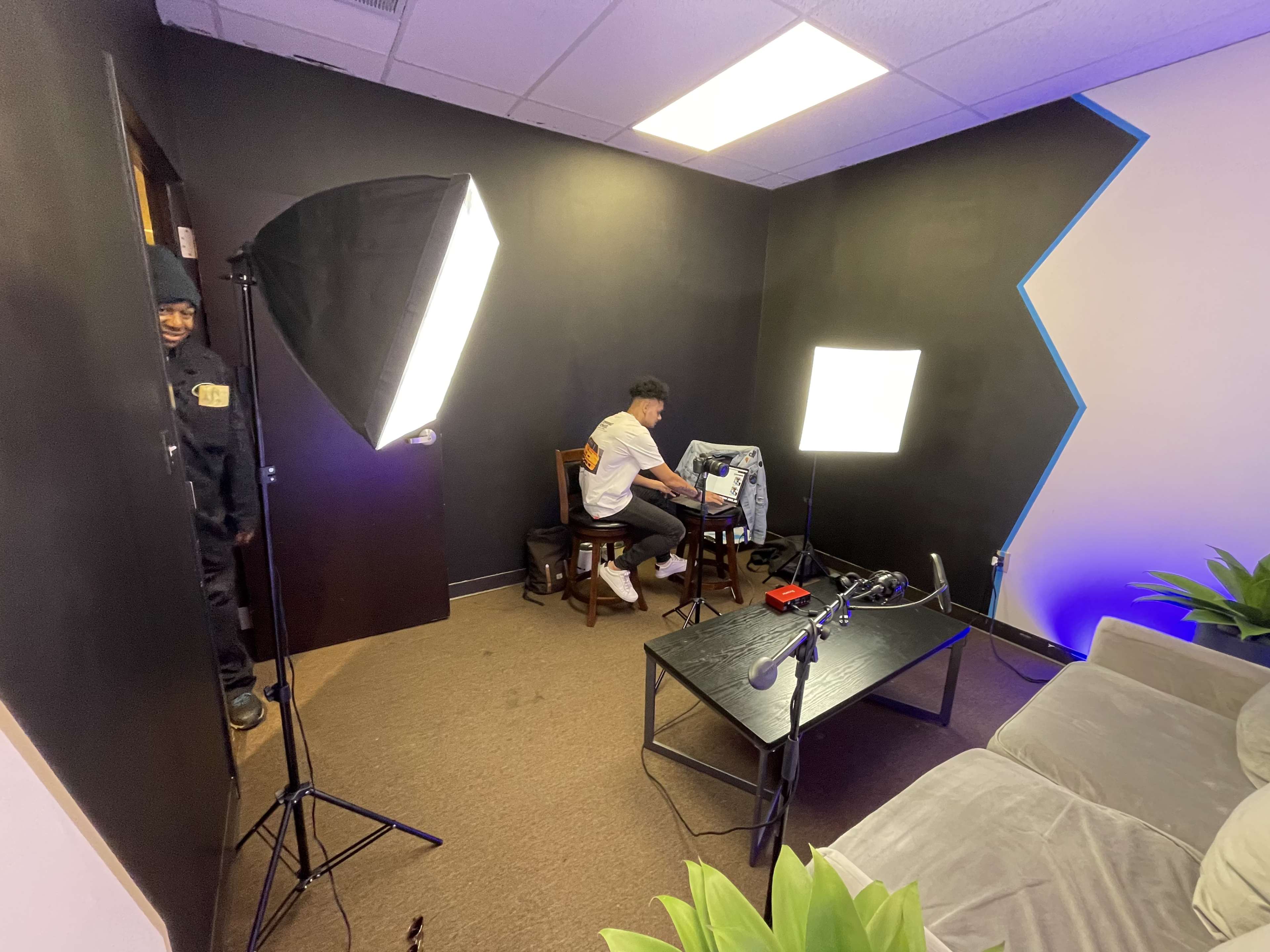 A man sits at a desk with a computer, surrounded by studio lighting and a microphone setup in a small room.
