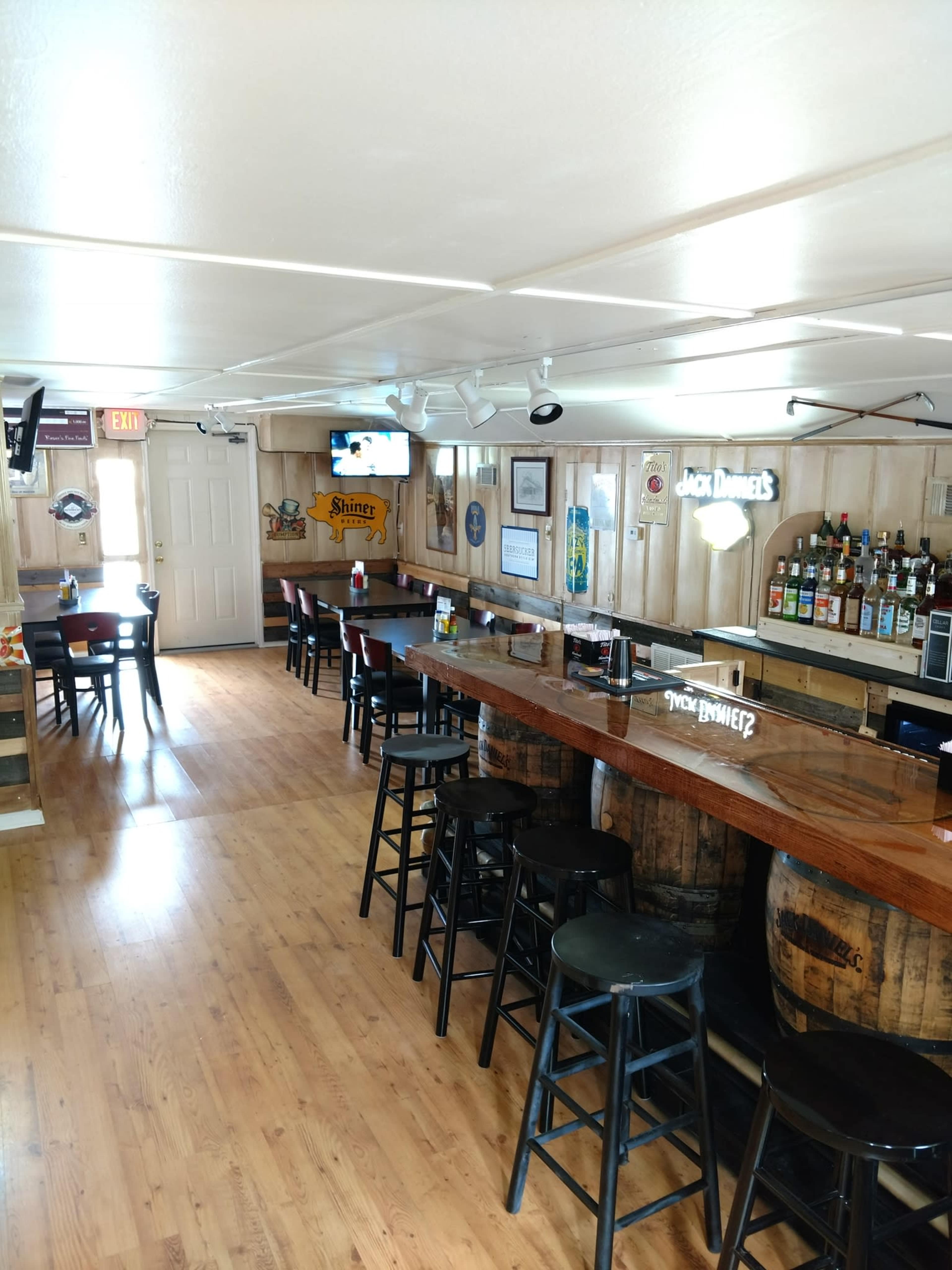 The image shows the interior of a bar with a wooden countertop, rows of black stools, and bottles displayed on shelves along the wall.