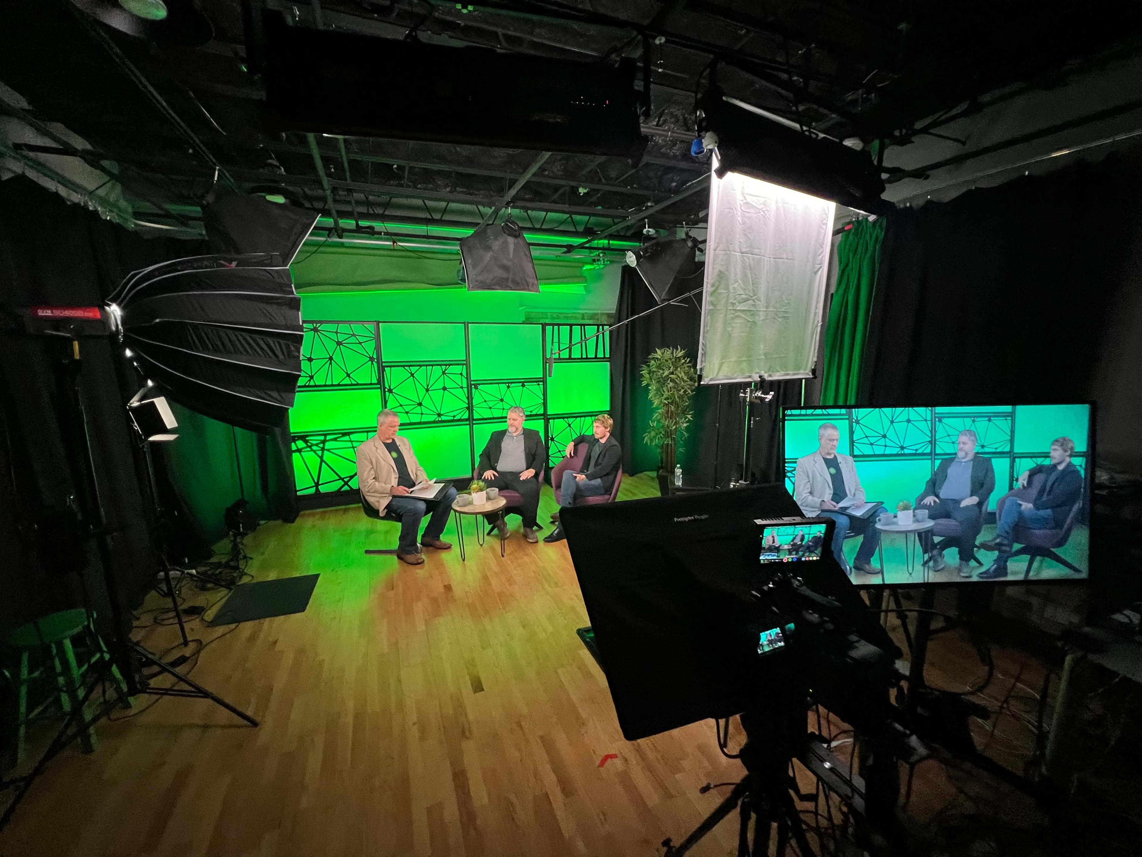 A television studio setup features three seated individuals engaging in discussion, with green backdrop lighting and multiple cameras positioned to capture the scene.